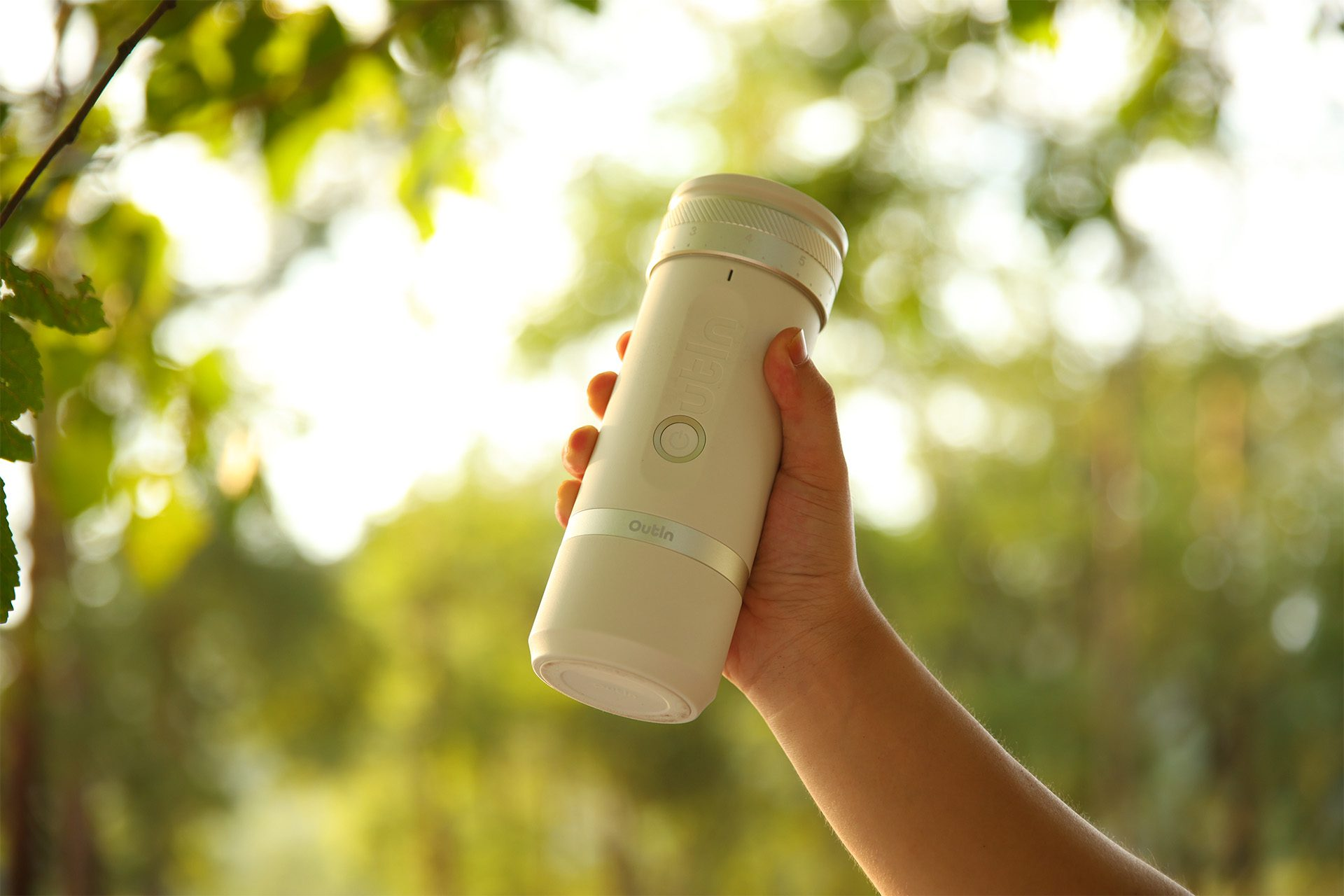 Dłoń trzyma białą butelkę Outin na rozmytym zielonym tle liści i światła słonecznego.