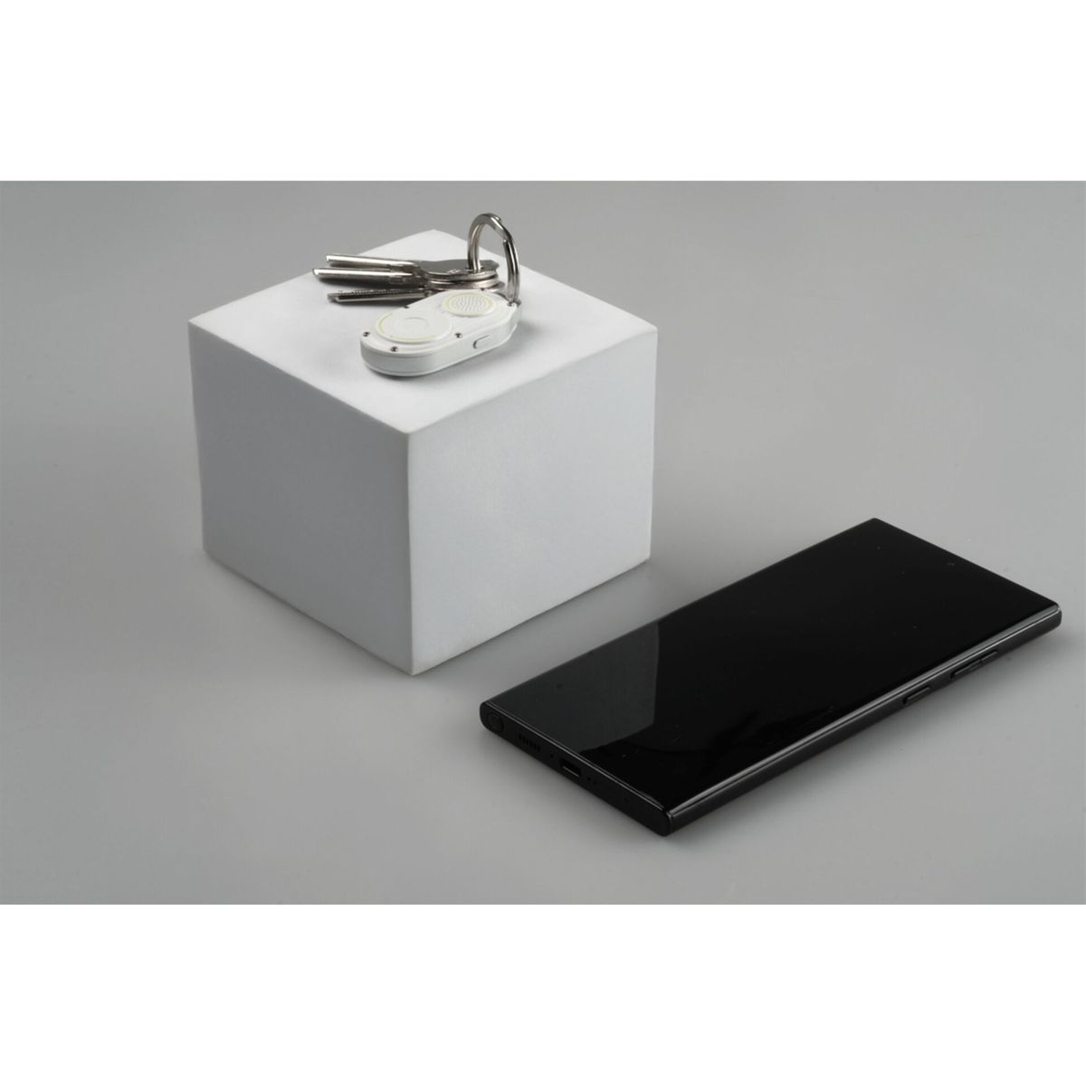 Een witte kubus met sleutels en een klein apparaat. Een zwarte smartphone ligt op een grijze ondergrond.