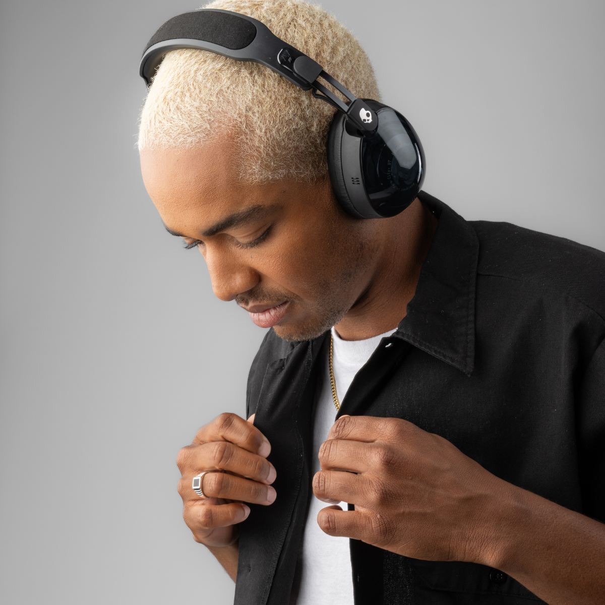 Mężczyzna z blond włosami w słuchawkach i czarnej koszuli, patrzący w dół. Ma pierścień i złoty łańcuch.