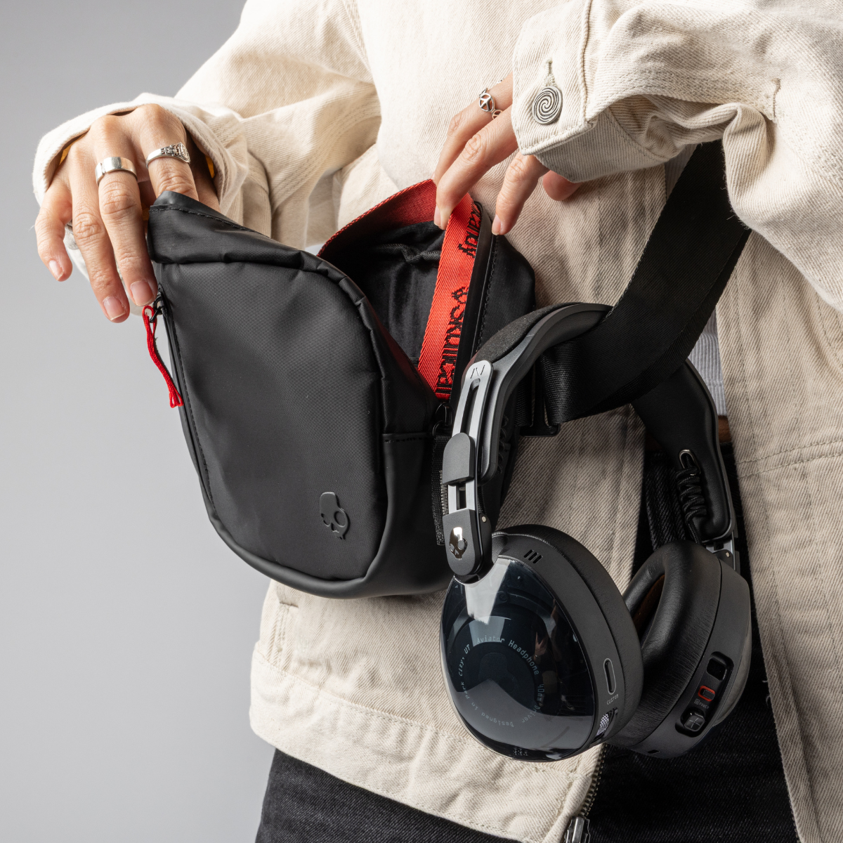 Osoba otwiera czarną torbę. Słuchawki wiszą na pasku torby. Osoba ma na sobie kremową kurtkę i pierścionki.