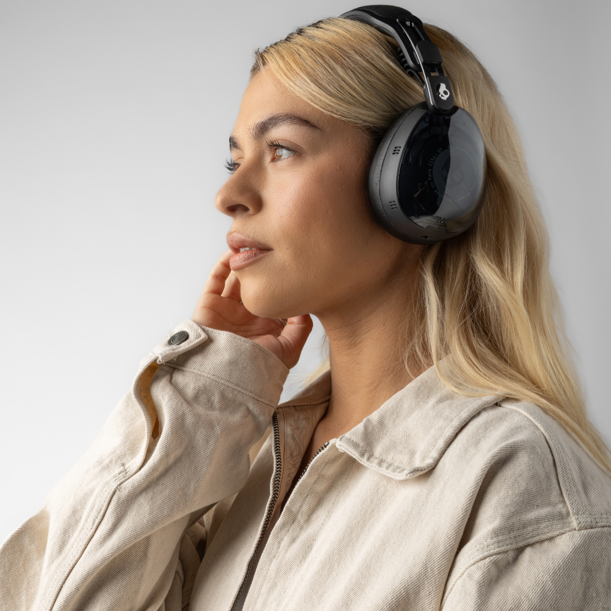 Blondynka w czarnych słuchawkach, beżowej kurtce, patrząca na bok, ręka dotyka twarzy, na białym tle.