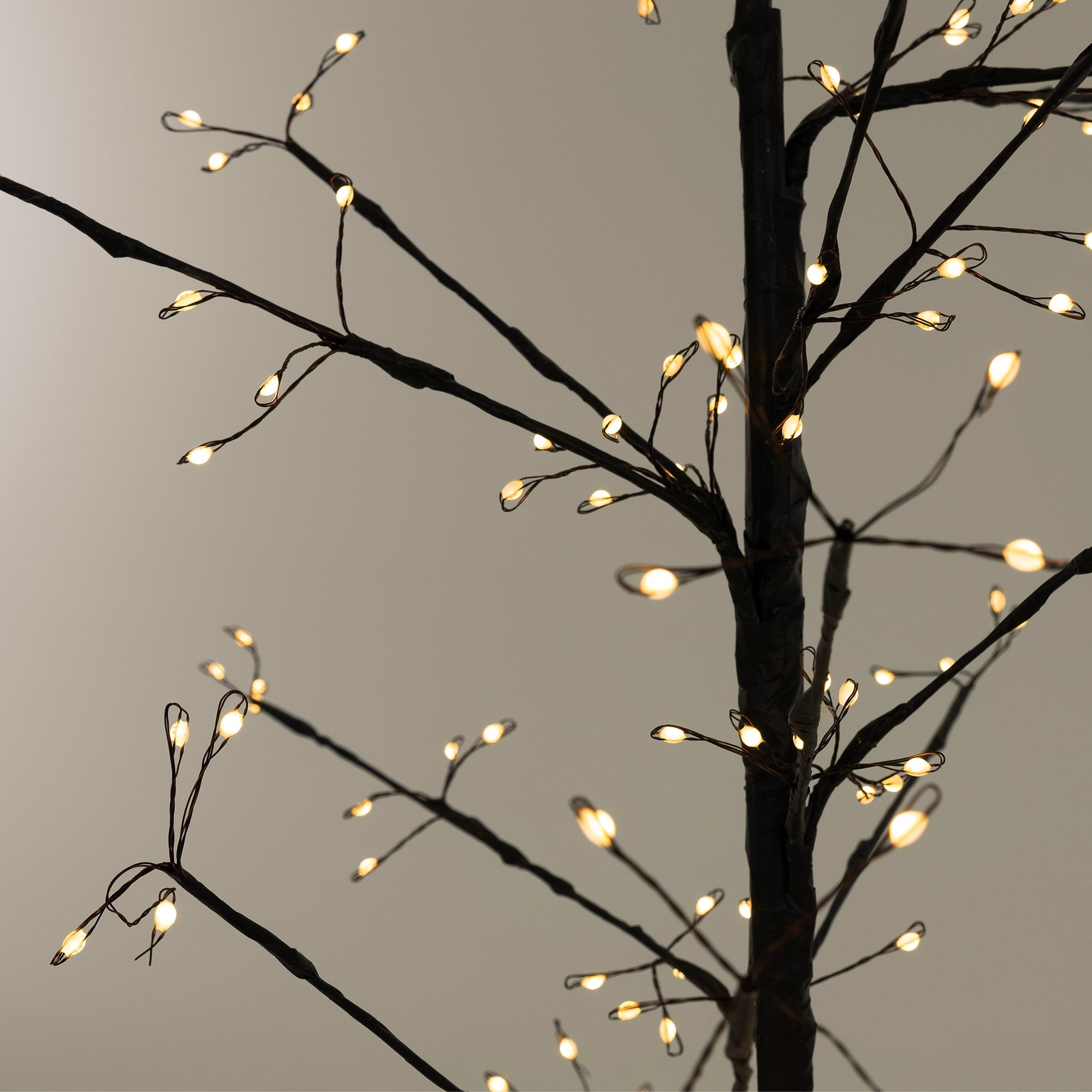 Nahaufnahme eines dekorativen Baumes mit vielen kleinen, warmweißen Lichtern auf schwarzen Ästen vor grauem Hintergrund.