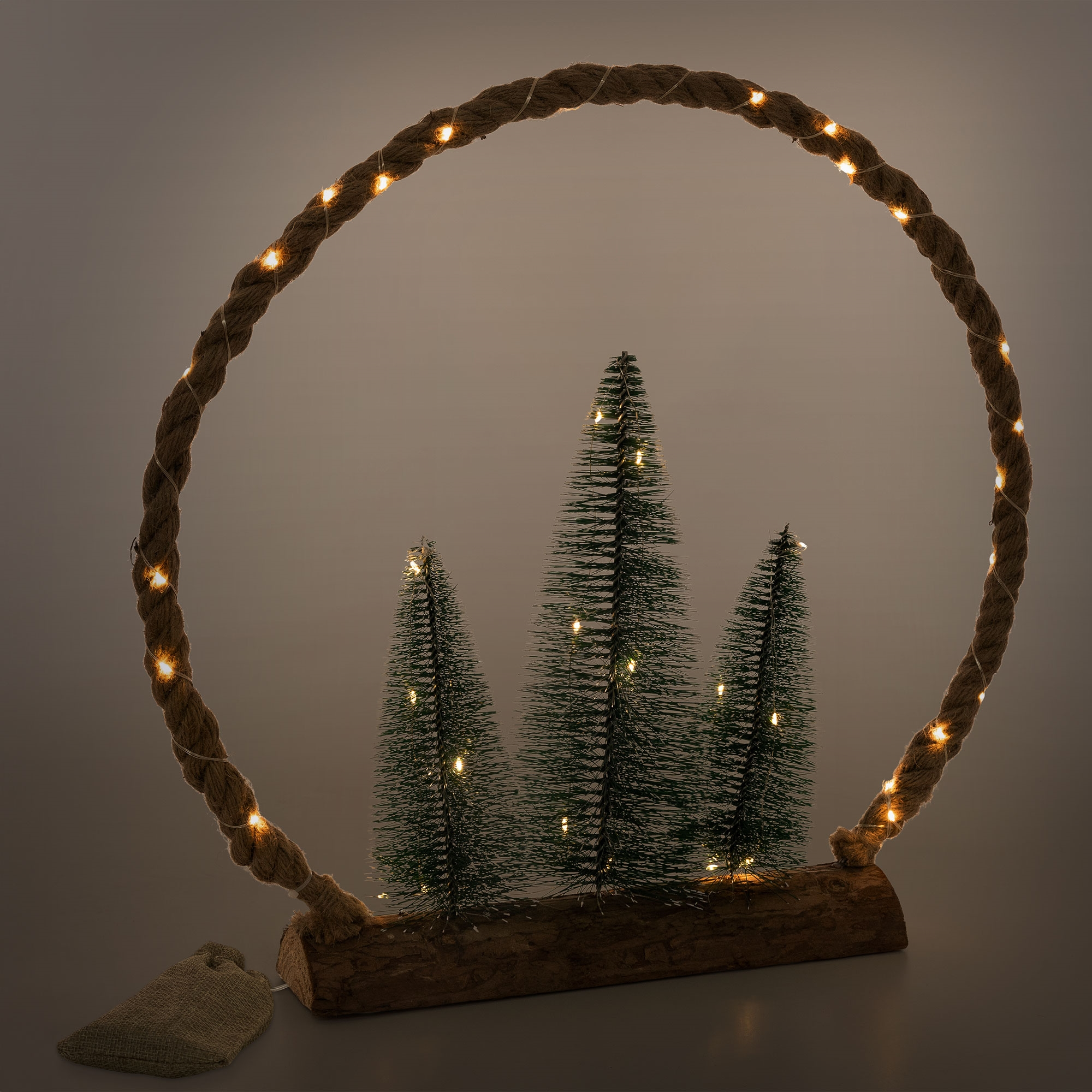 Eine Weihnachtsdekoration mit drei kleinen Tannenbäumen in einem Seilkreis mit Lichtern, auf einem Holzsockel, mit einem Jutesack.