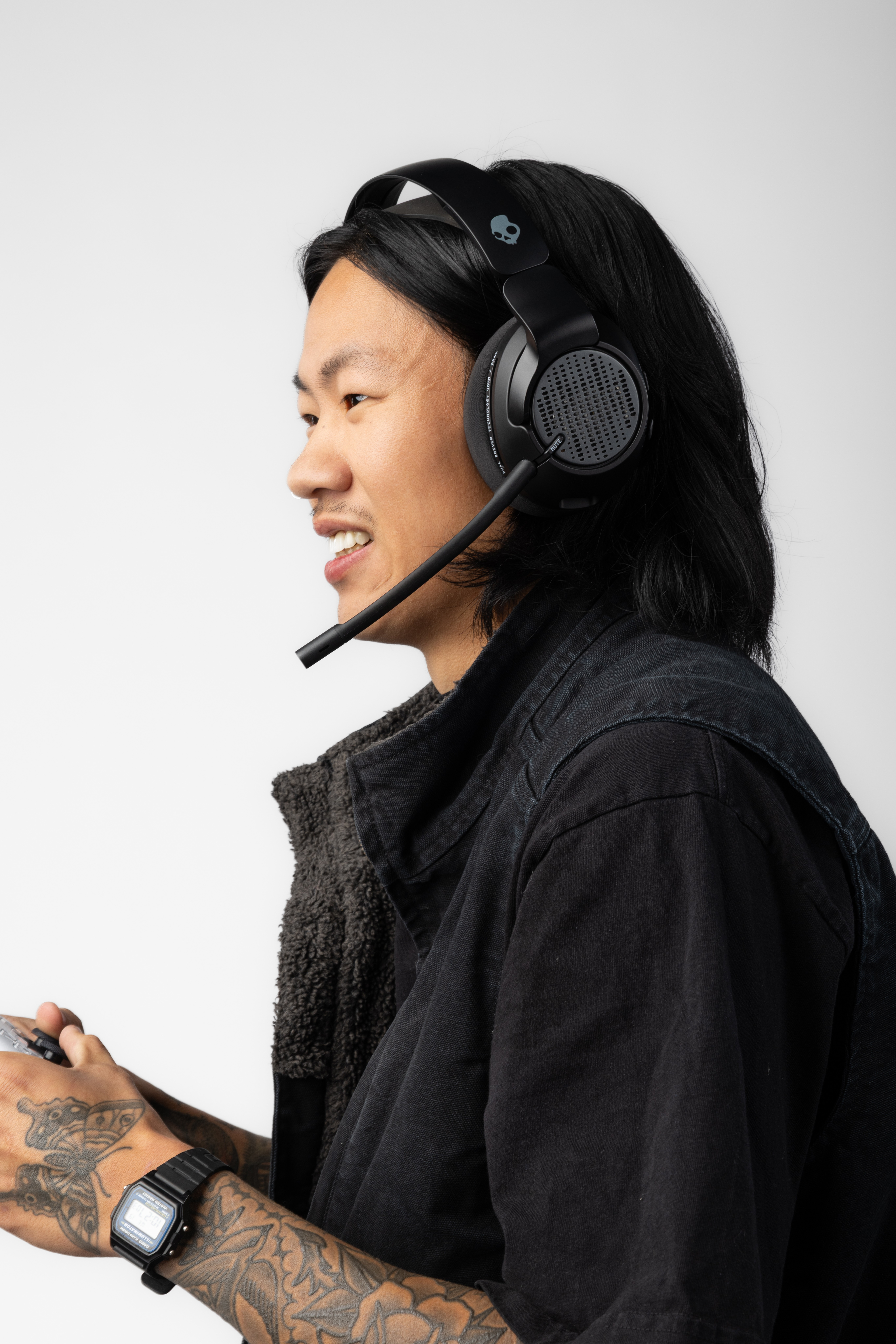 Mężczyzna w słuchawkach i zestawie słuchawkowym grający. Ma na sobie czarną kurtkę i tatuaże. Białe tło.