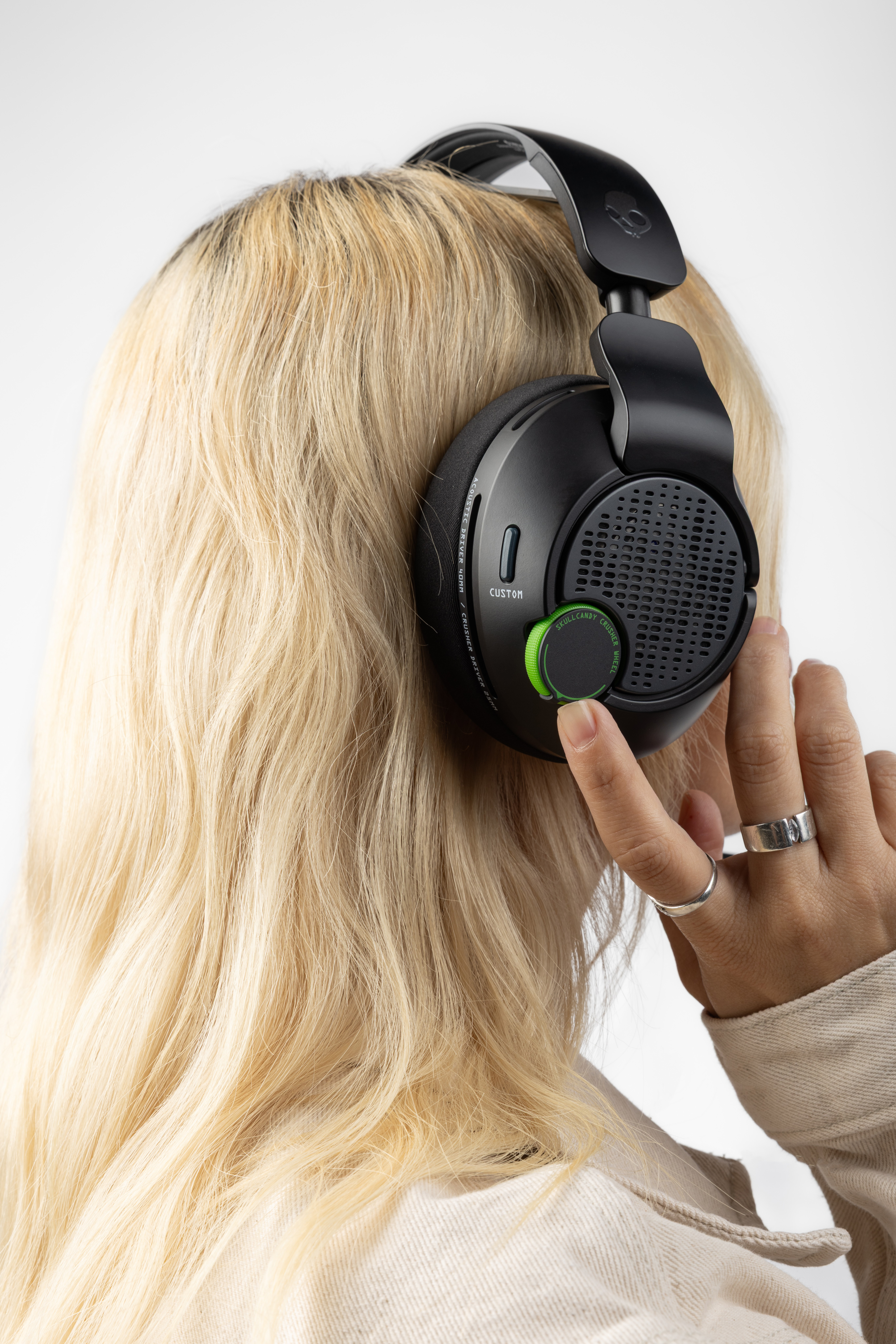 Osoba z blond włosami nosi czarne słuchawki. Ręka reguluje zielone pokrętło. Palce zdobią pierścienie.