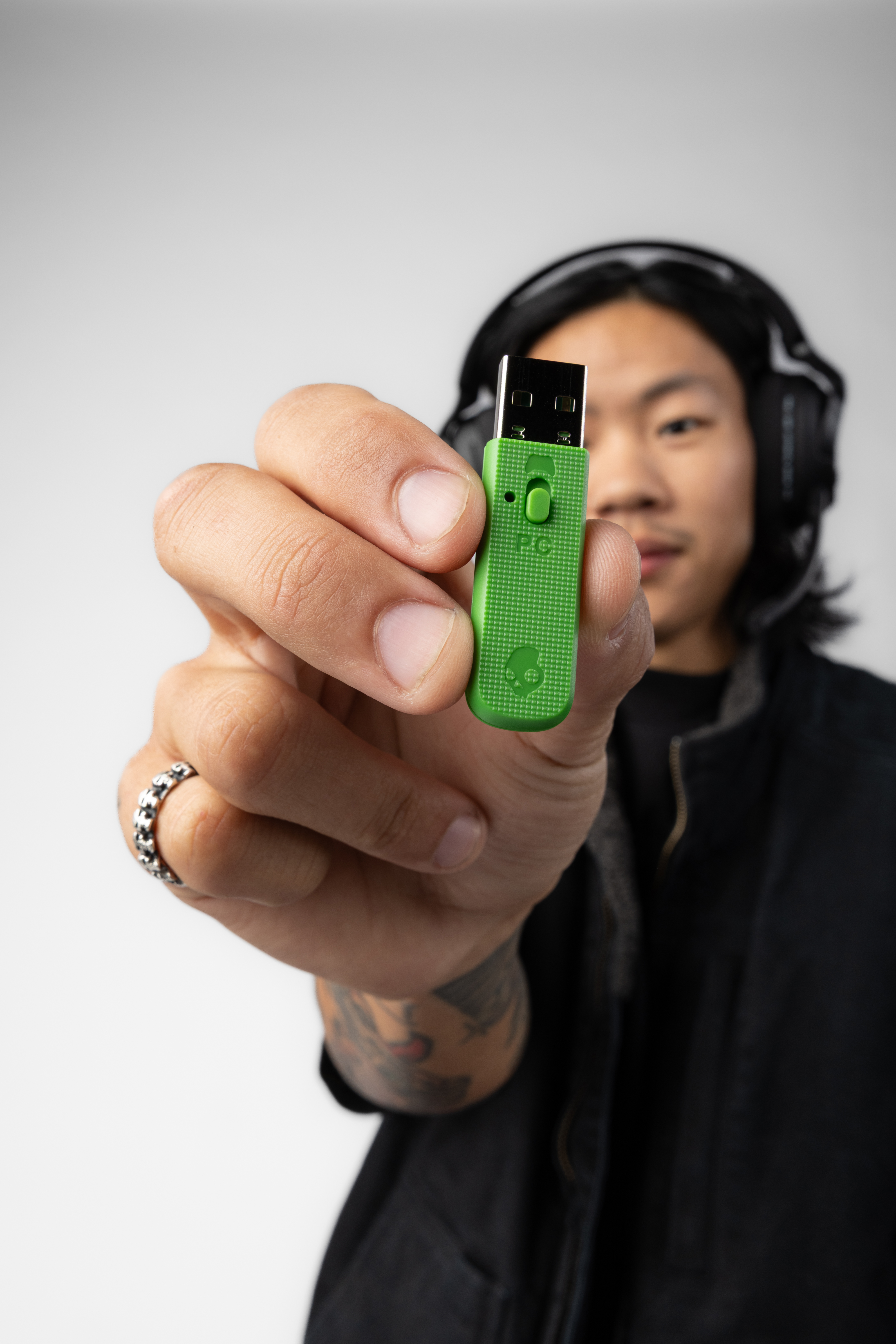 Osoba trzyma zielony klucz USB. Osoba ma na sobie słuchawki i czarną kurtkę. Białe tło.