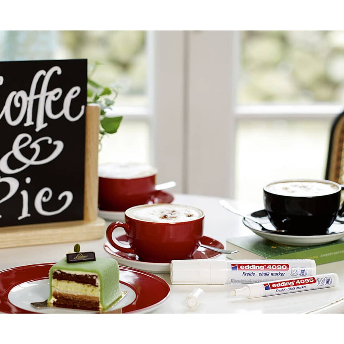 Szene in einem Café. Rote Kaffeetassen, ein Kuchen und Kreidemarker stehen auf einem weißen Tisch. Ein Schild sagt 'Kaffee & Kuchen'.