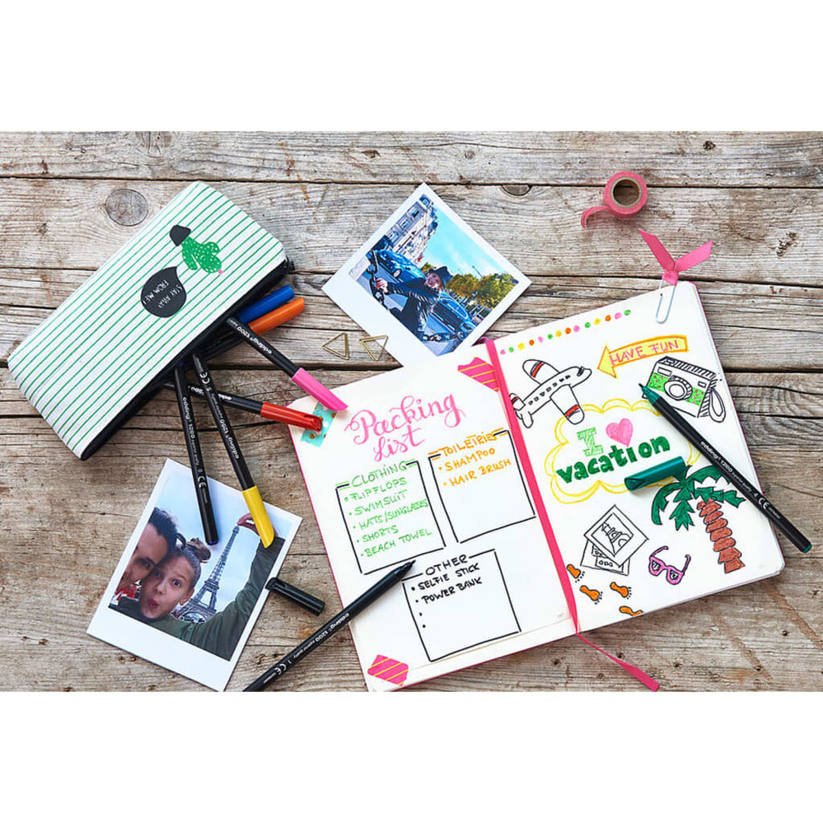 Eine Packliste mit Zeichnungen und Fotos auf einer Holzoberfläche, Stiften, einem Federmäppchen und Klebeband. Das Thema ist Urlaub.