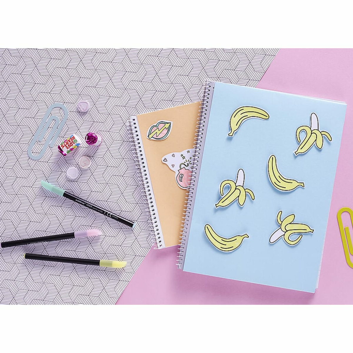 Notizbücher mit Bananenaufklebern, Stiften, Büroklammern und Süßigkeiten liegen auf einer gemusterten Oberfläche, auf rosa Hintergrund.