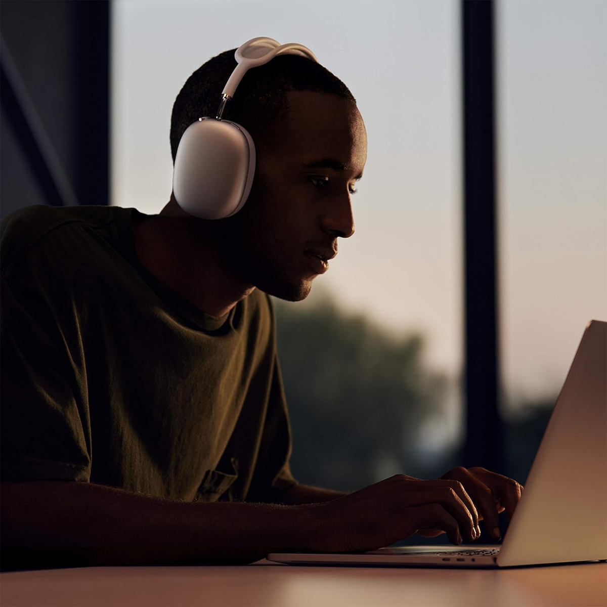 Ein Mann mit Kopfhörern tippt auf einem Laptop. Er trägt ein grünes Hemd, sitzt an einem Tisch und hat ein Fenster im Hintergrund.
