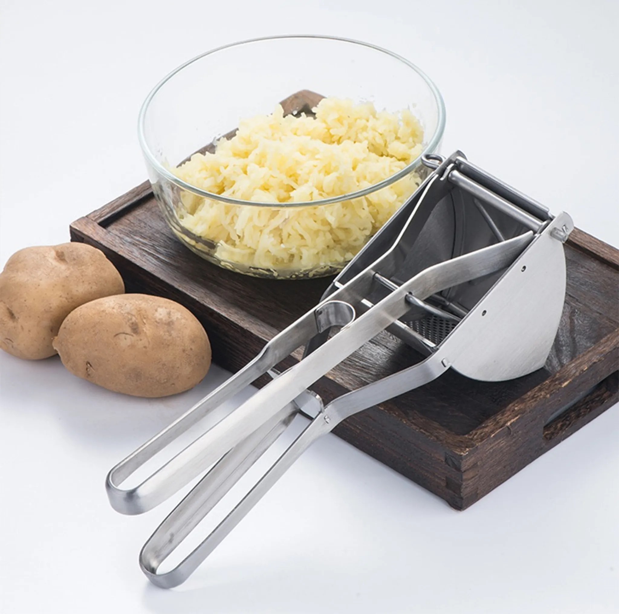 Kartoffelpresse, Schüssel mit Kartoffelreis und zwei ganze Kartoffeln auf einem Holztablett. Weißer Hintergrund.