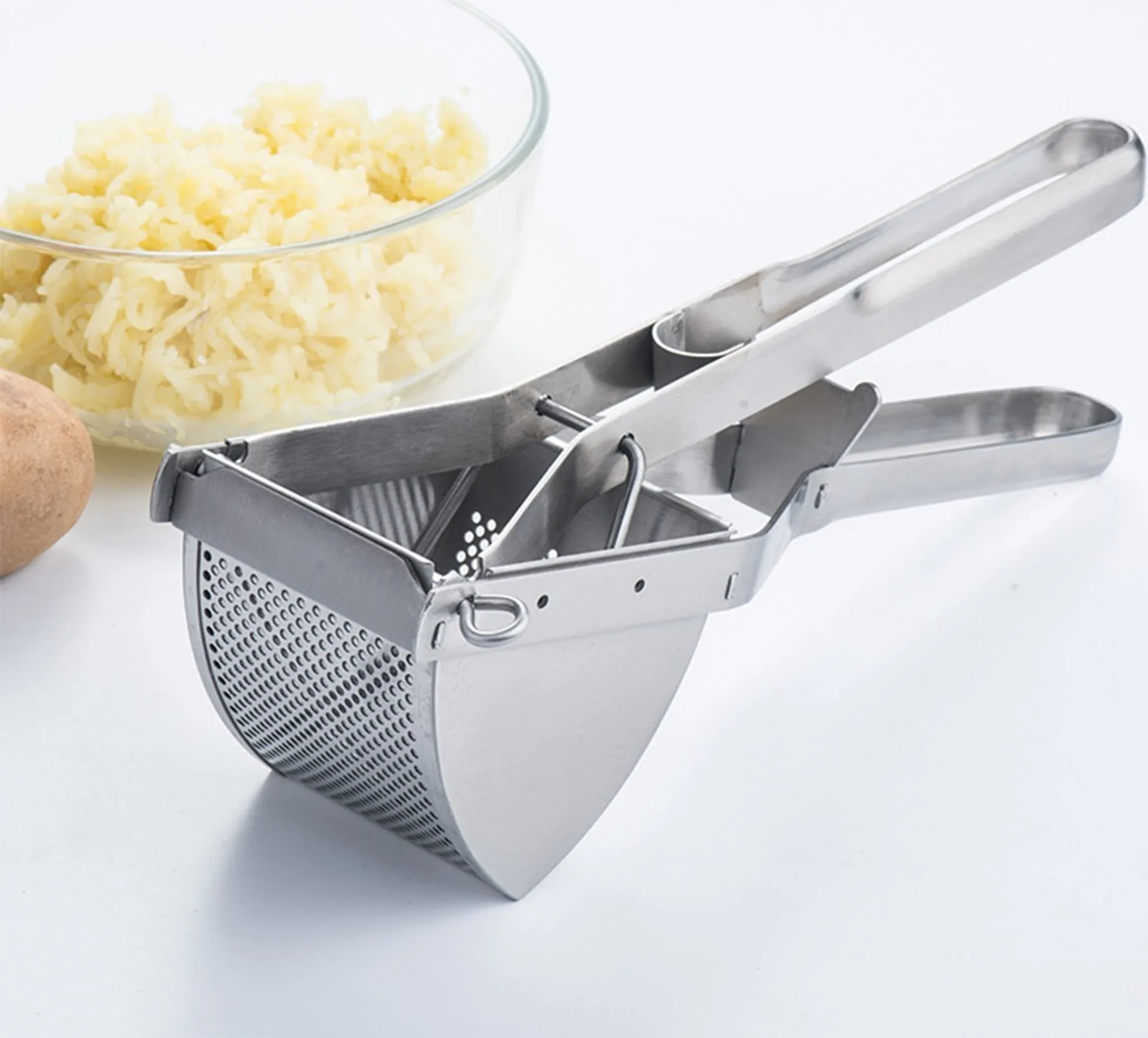 Eine Kartoffelpresse aus Edelstahl mit Griffen, neben einer Schüssel mit Kartoffelpüree und einer Kartoffel.