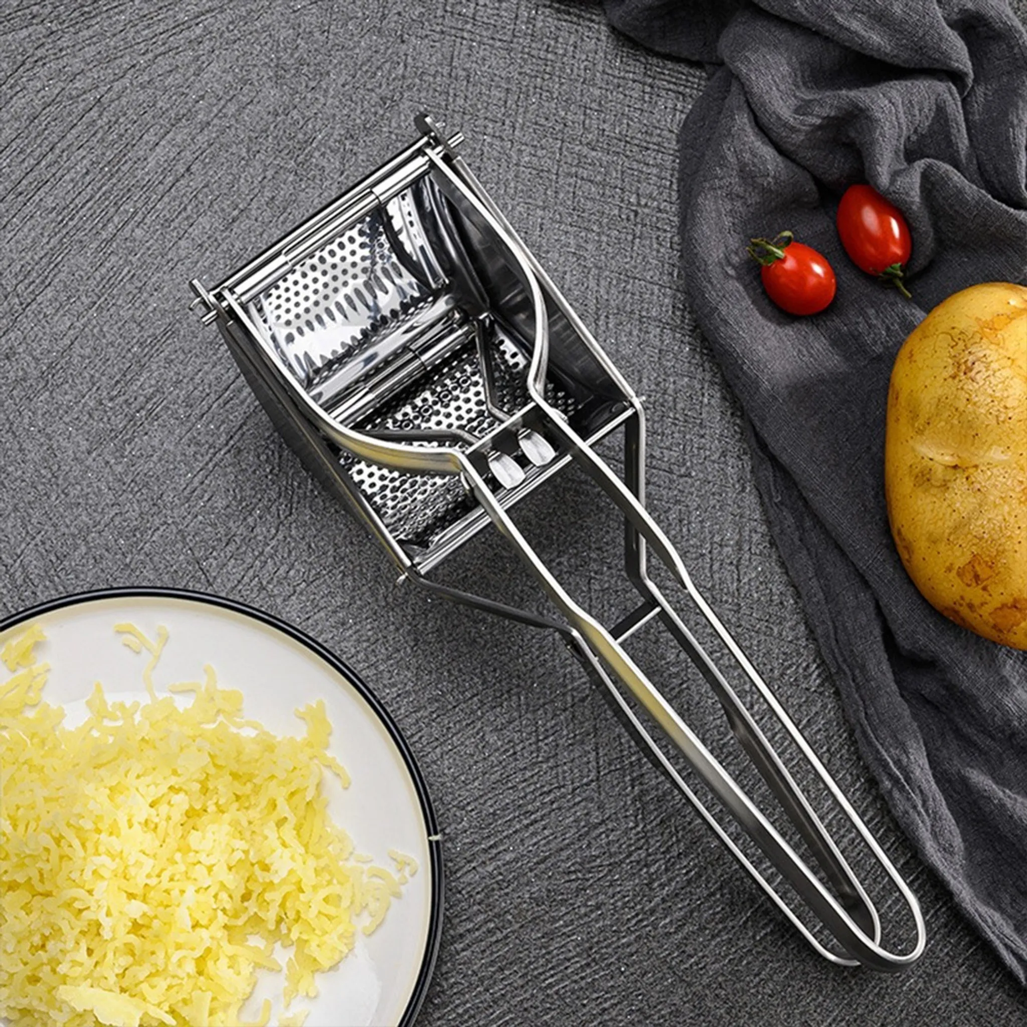 Ein Kartoffelstampfer, Kartoffelpüree, Tomaten und eine Kartoffel liegen auf einer grauen Oberfläche. Auf der linken Seite befindet sich ein Teller mit geriebenen Kartoffeln.