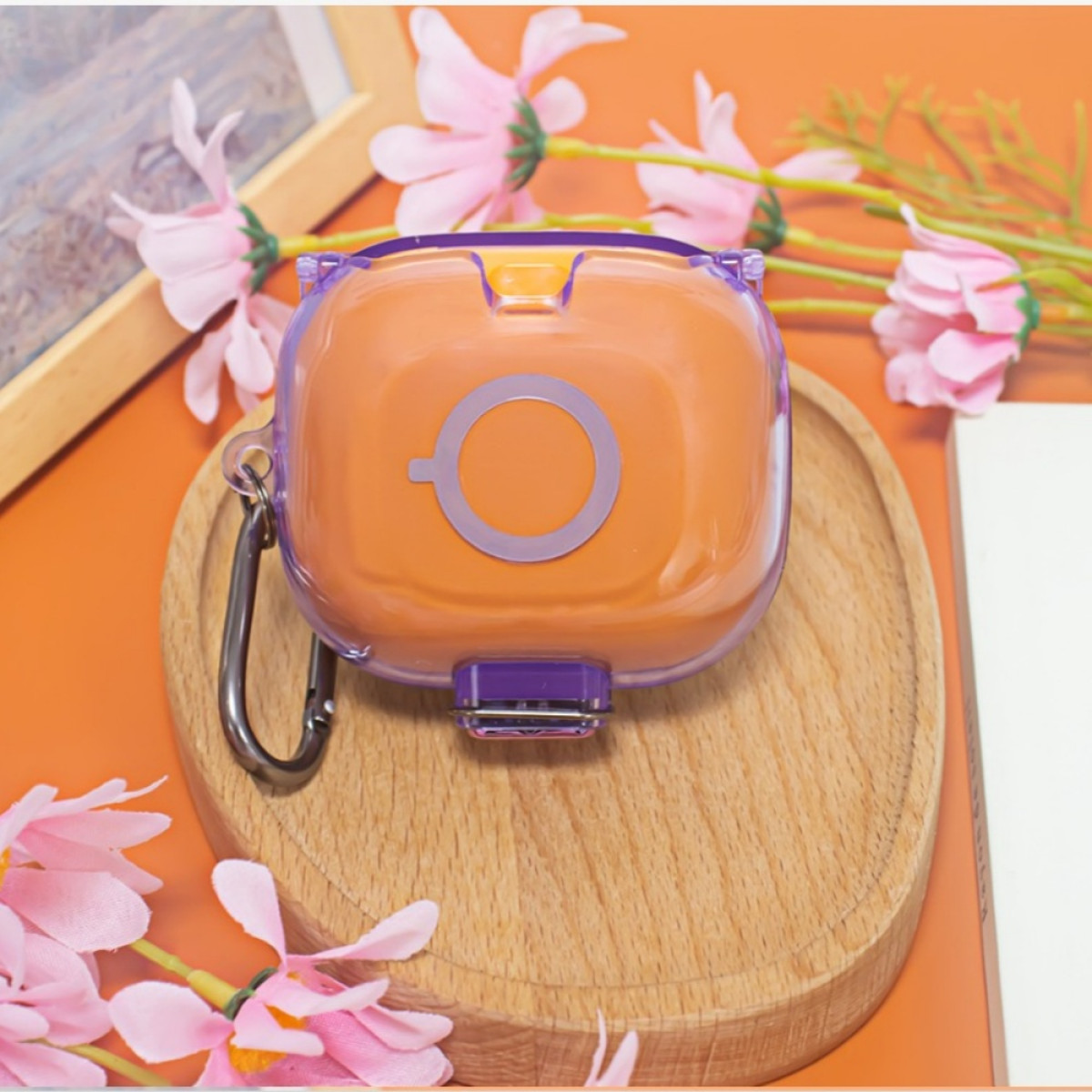 Ein orangefarbenes Etui mit lila Akzenten liegt auf einem Holzbrett. Rosa Blumen und ein Rahmen befinden sich im Hintergrund. Der Hintergrund ist orange.