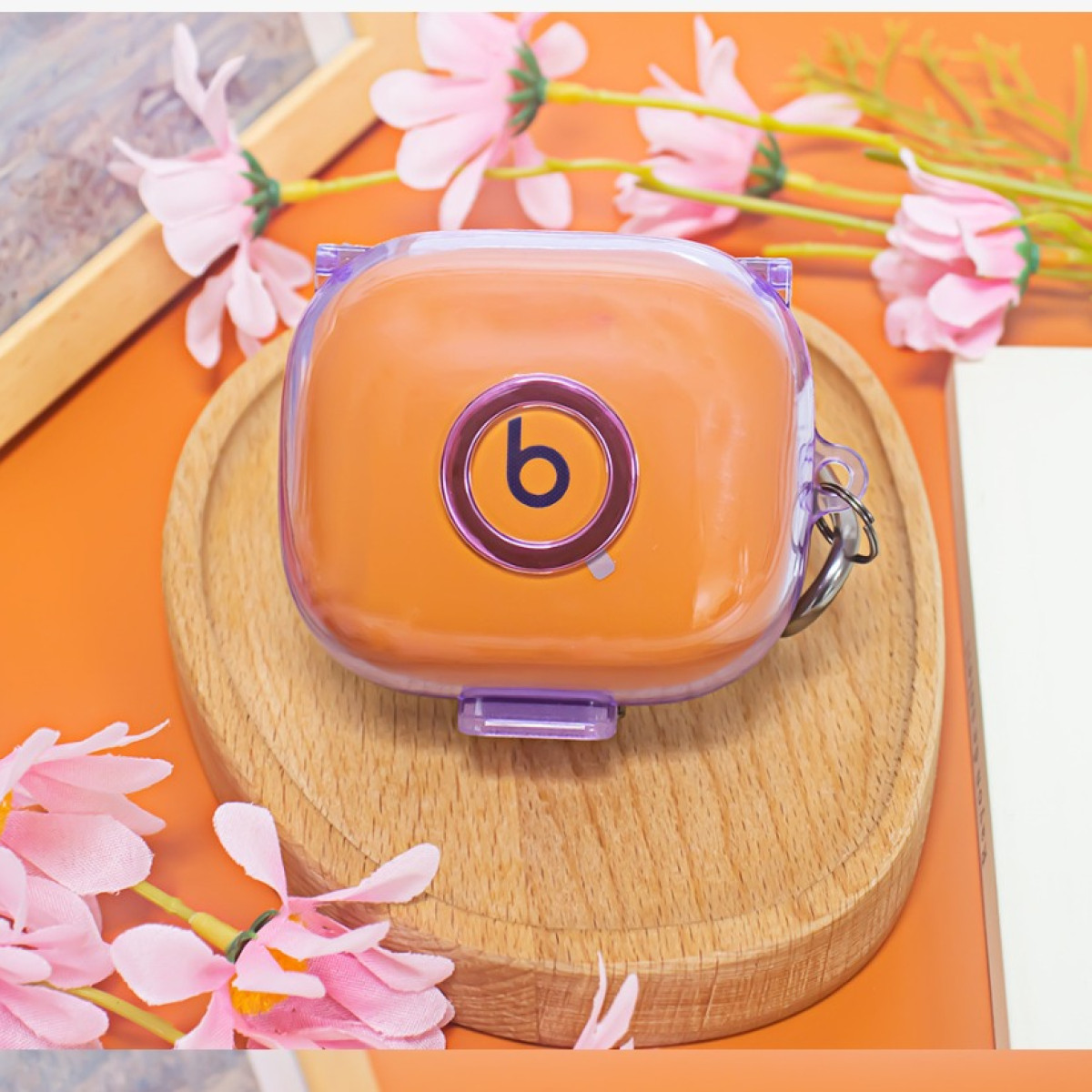 Orangefarbene Beats-Kopfhörerhülle auf Holztablett, umgeben von rosa Blumen. Die Hülle hat einen lilafarbenen Ring mit dem 'b'-Logo.