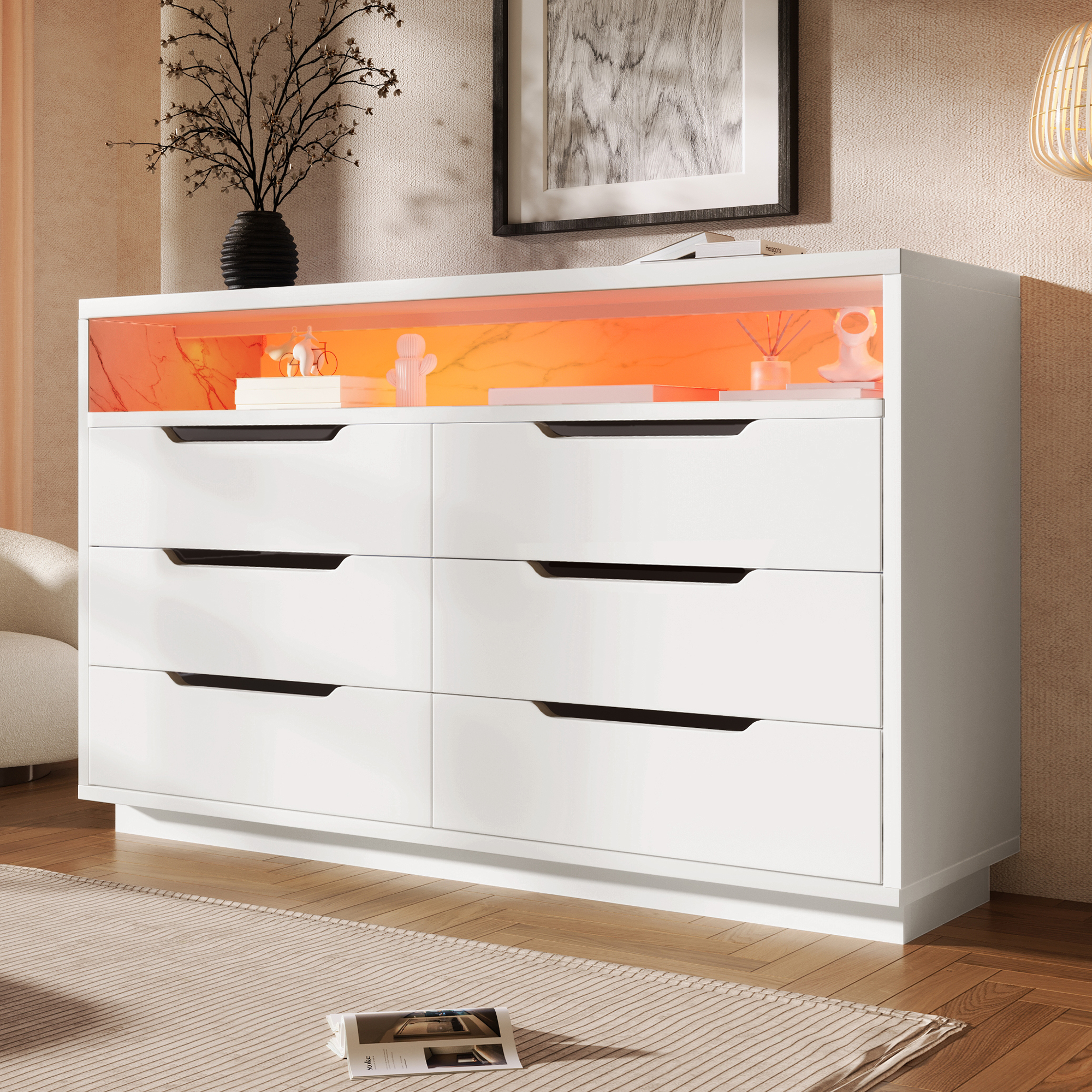 Eine weiße Kommode mit sechs Schubladen. Darüber befindet sich ein beleuchtetes Regal mit Dekorationen. Eine Vase mit Zweigen steht auf der Kommode.