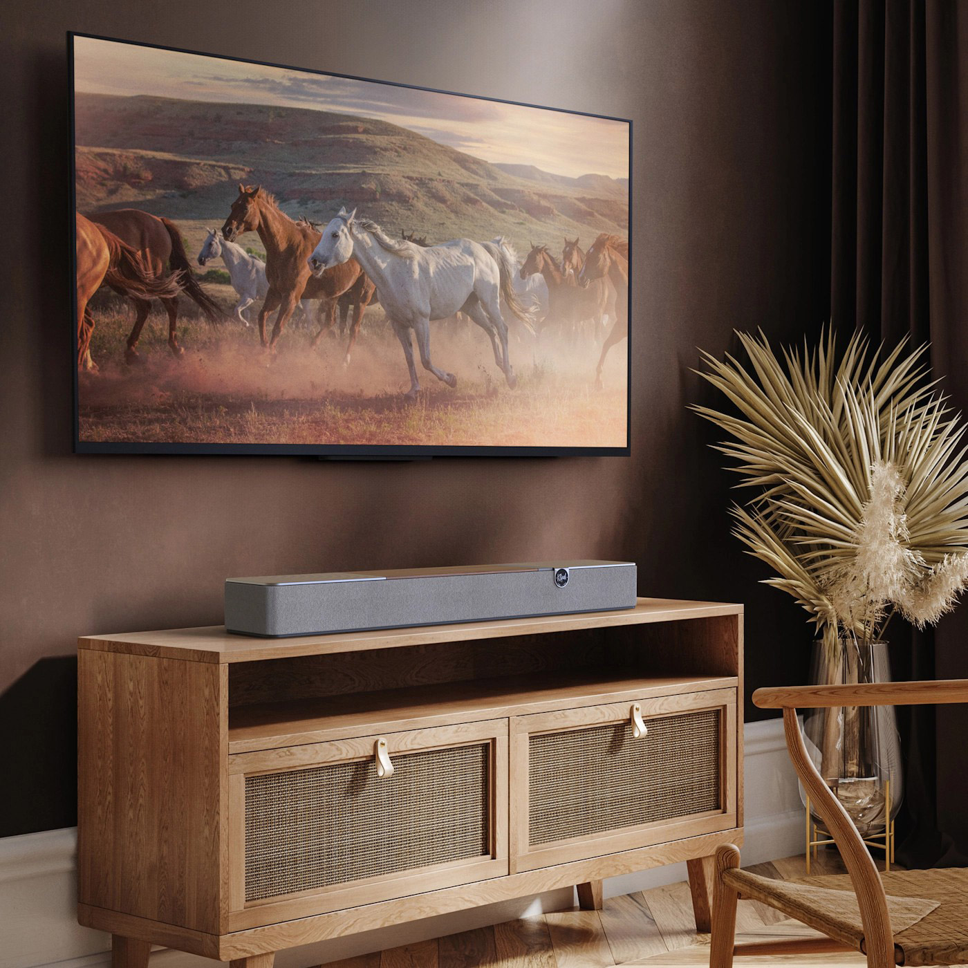 Un televisore mostra cavalli che corrono. Una soundbar è su un mobile di legno. Una sedia e un vaso con piante secche sono vicini.