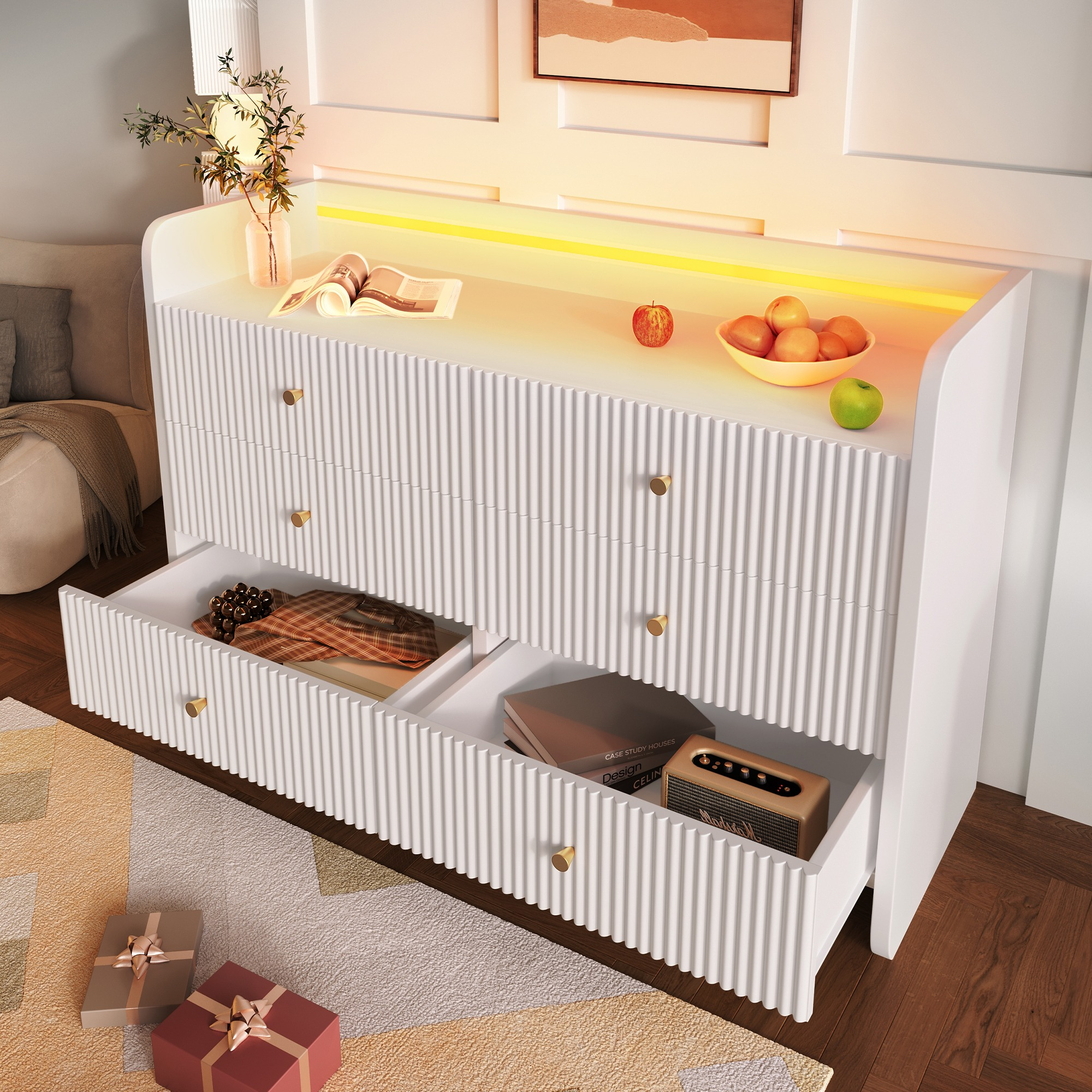 Weißer Schrank mit offenen Schubladen, Obstschale, Büchern und Geschenken auf dem Boden. Eine Vase mit Pflanzen steht oben.