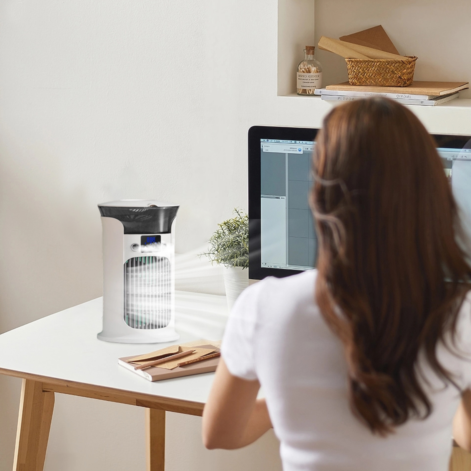 Eine Frau sitzt an einem Schreibtisch und arbeitet an einem Computer. Auf dem Schreibtisch steht ein weißer Luftkühler. Der Hintergrund hat ein Regal mit Dekoration.