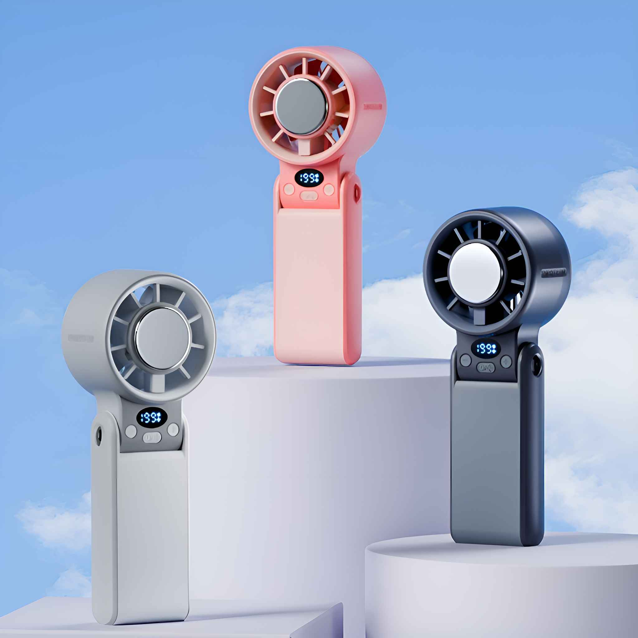 Drei tragbare Ventilatoren in Rosa, Grau und Dunkelgrau, vor blauem Himmel und weißem Podest.