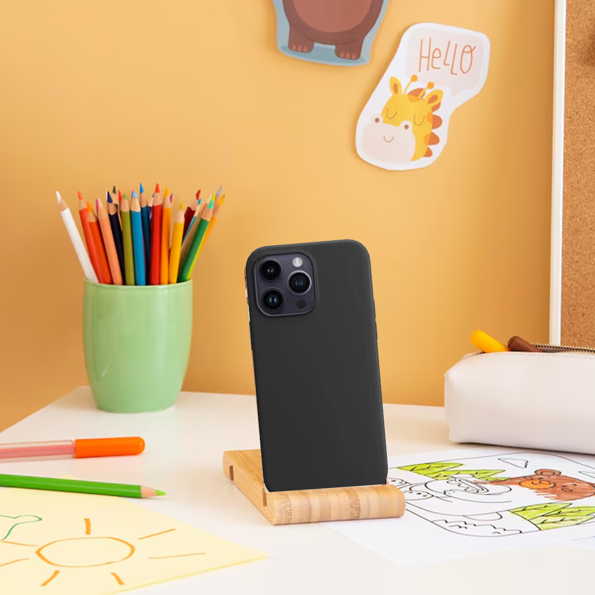 Ein schwarzes Smartphone auf einem Bambusständer steht auf einem weißen Schreibtisch. Buntstifte in einem grünen Becher, Zeichnungen und Aufkleber sind sichtbar.