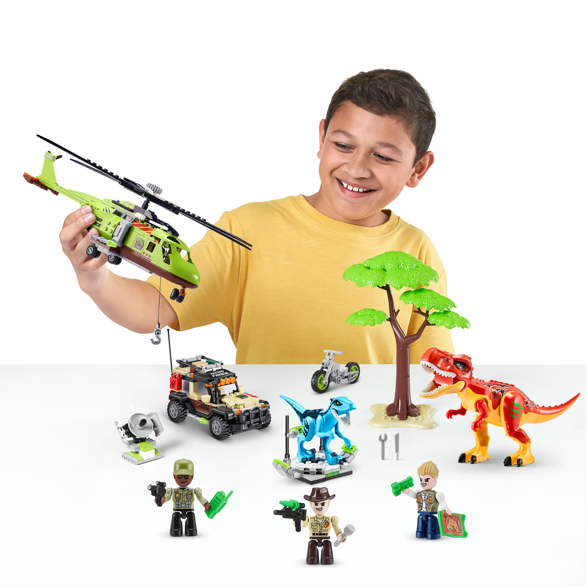 Chłopiec trzyma zielony helikopter. Zabawkowe dinozaury, pojazdy i figurki ułożone na białej powierzchni.