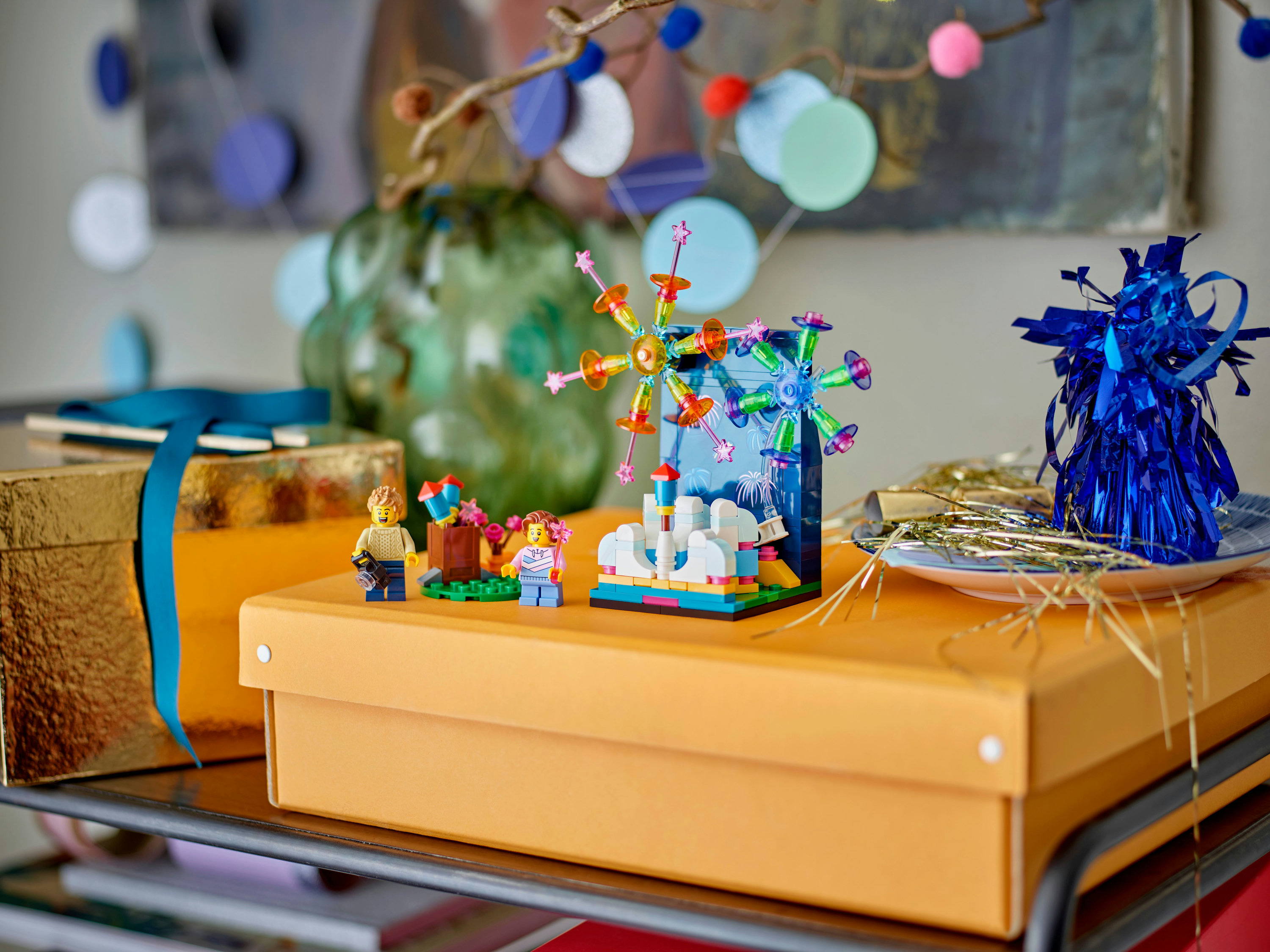 Eine orangefarbene Schachtel mit Lego-Figuren und einem Riesenrad. Blaue Schleife, Partydekorationen und ein grünes Objekt im Hintergrund.