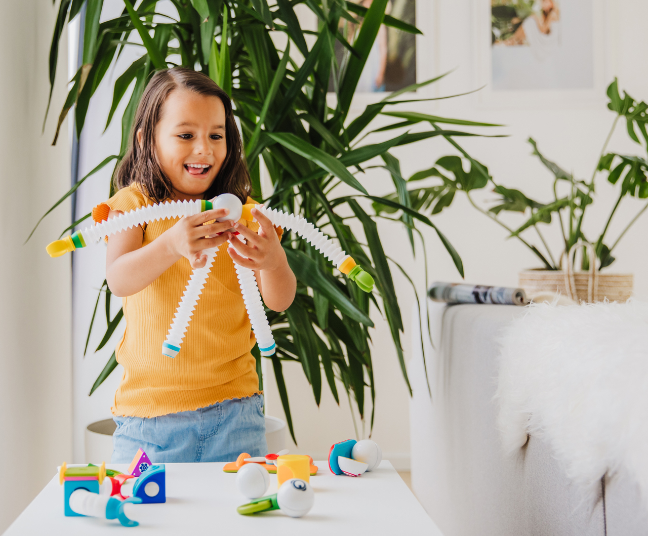 Una bambina sorridente con una maglietta gialla gioca con un giocattolo bianco multi-articolato. Giocattoli su un tavolo bianco. Piante verdi sullo sfondo.