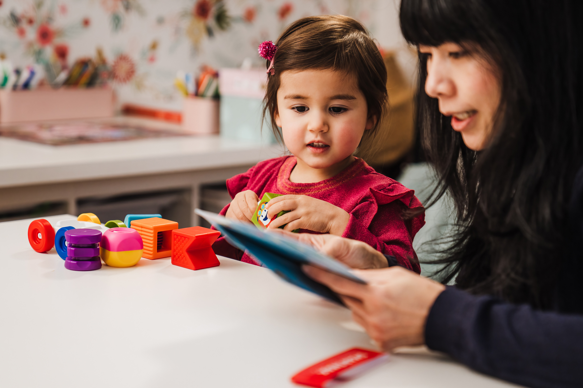 Una bambina e un adulto sono seduti a un tavolo. La bambina tiene in mano un giocattolo e guarda un libro. Ci sono blocchi colorati sul tavolo.