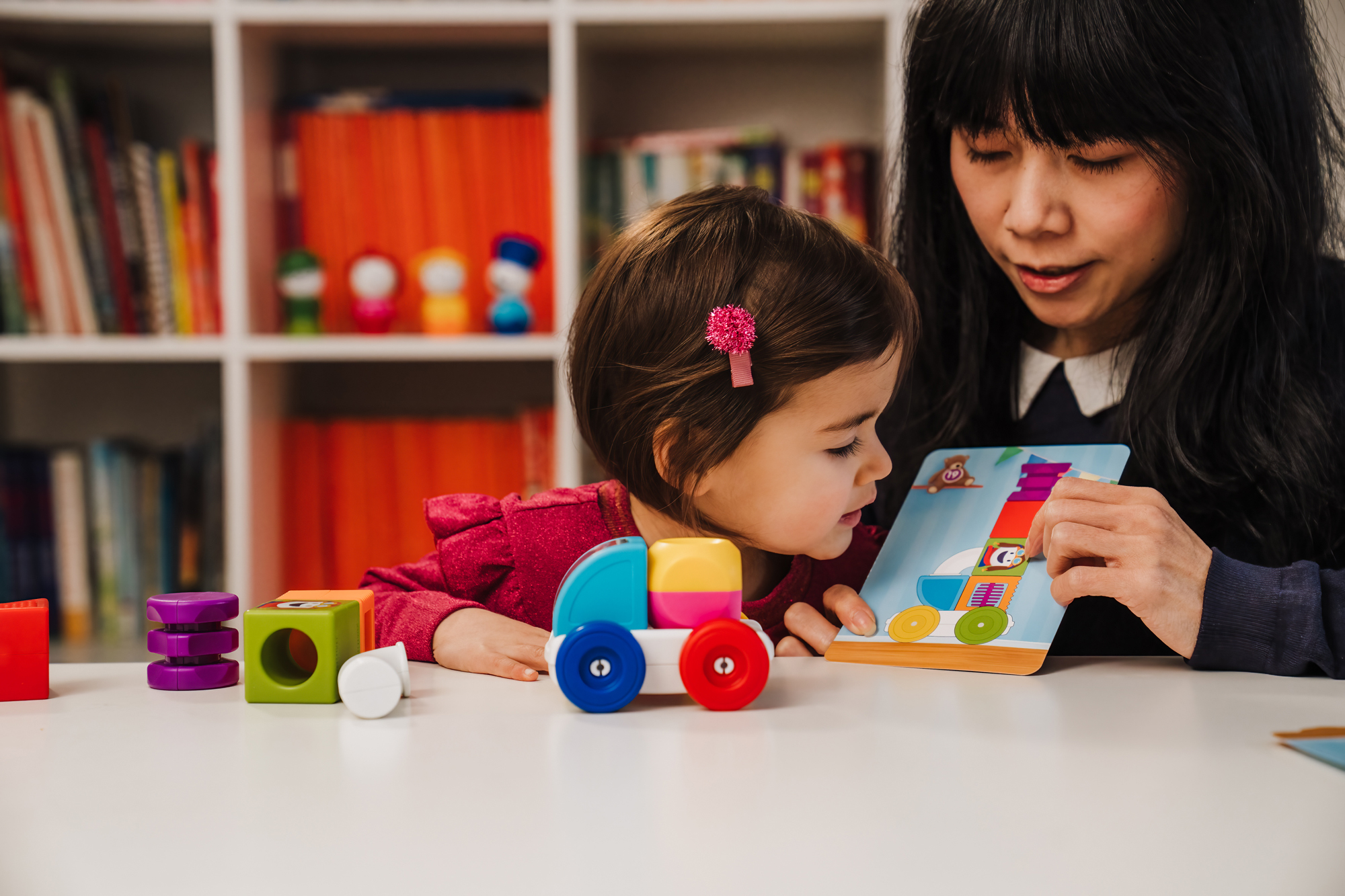 Una donna e un bambino guardano un libro. I giocattoli sono sul tavolo. Librerie sullo sfondo. La scena è luminosa.