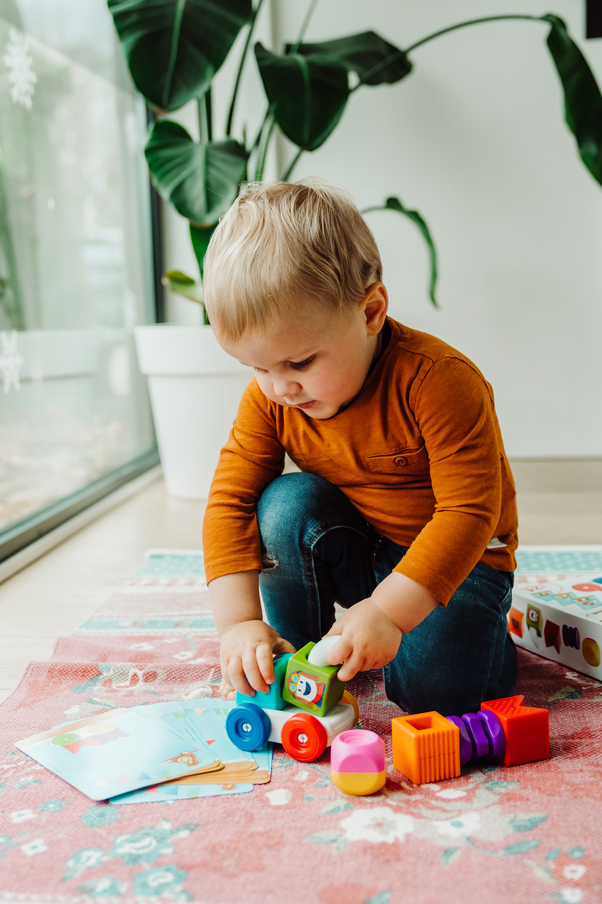 Un bambino gioca con blocchi colorati su un tappeto. Il bambino indossa una camicia arancione e jeans blu.