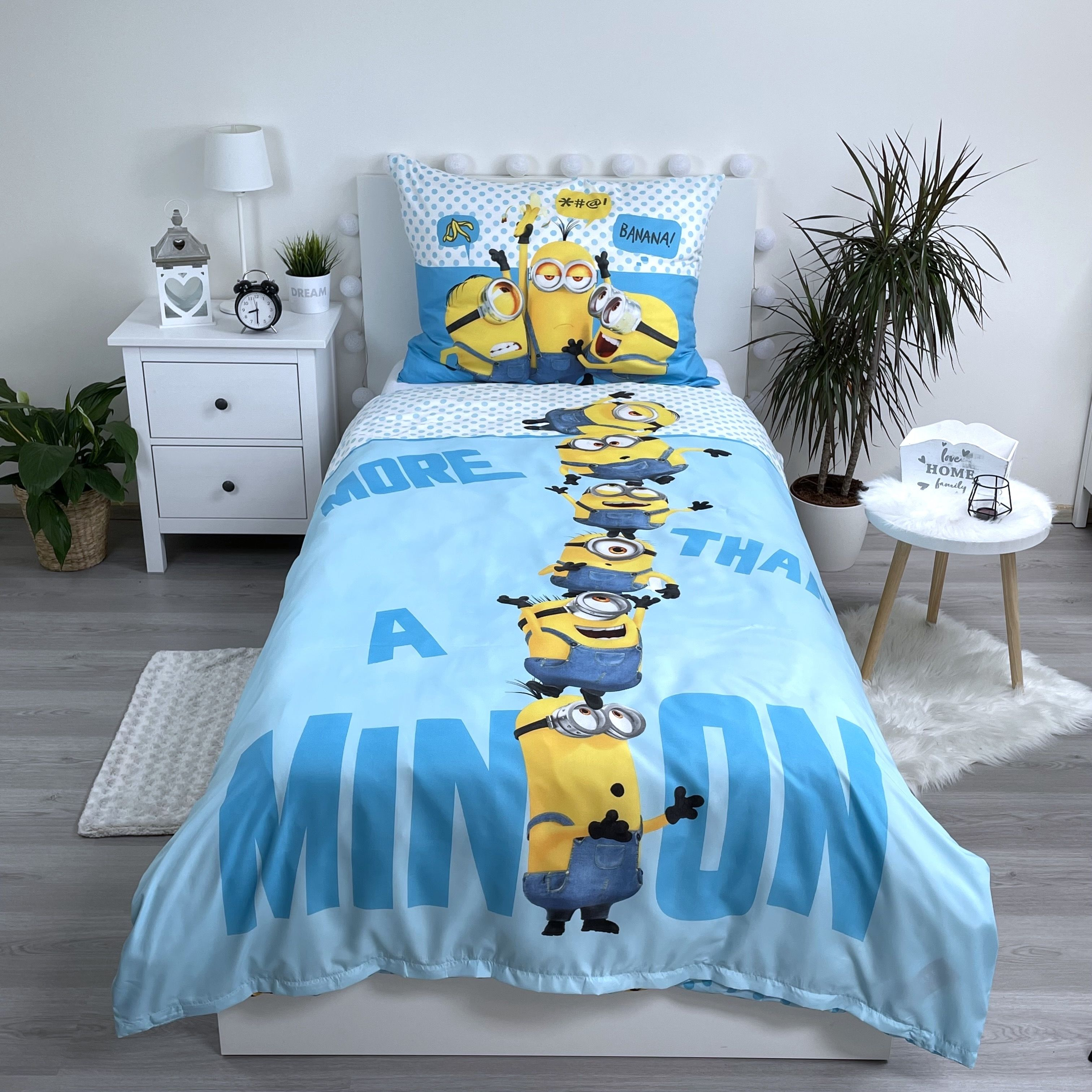 Ein Bett mit Minions-Bettwäsche. Ein weißer Nachttisch hat eine Lampe, einen Wecker und eine Pflanze. Auf dem Boden steht eine Pflanze.