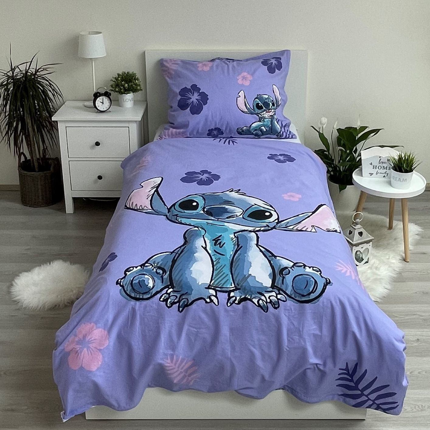 Bettwäsche im Stitch-Design. Lila Bettwäsche mit Stitch-Grafik. Nachttisch mit Lampe und Uhr. Pflanzen und ein kleiner Teppich.