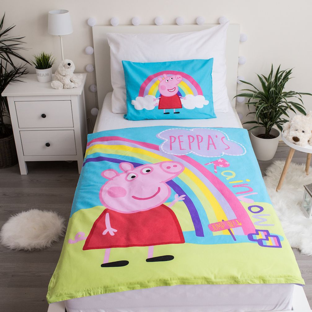 Ein Bett mit einer Peppa Wutz-Bettwäsche und einem Kissenbezug mit einem Regenbogen. Ein kleiner weißer Nachttisch mit Pflanzen ist sichtbar.