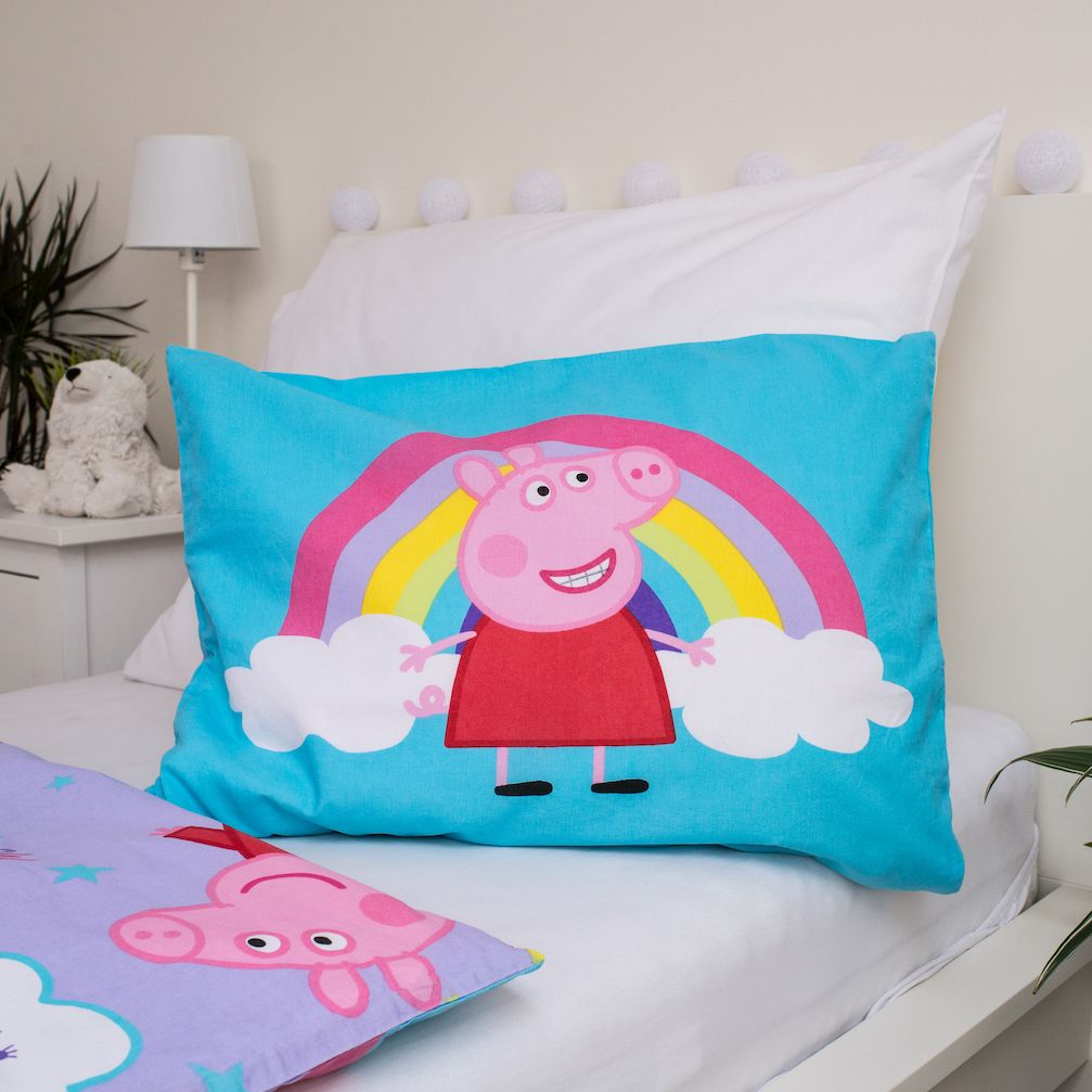 Blaues Kissen mit Peppa Wutz und Regenbogen-Design. Es liegt auf einem Bett mit einem anderen Kissen. Weiße Bettwäsche und ein Nachttisch.