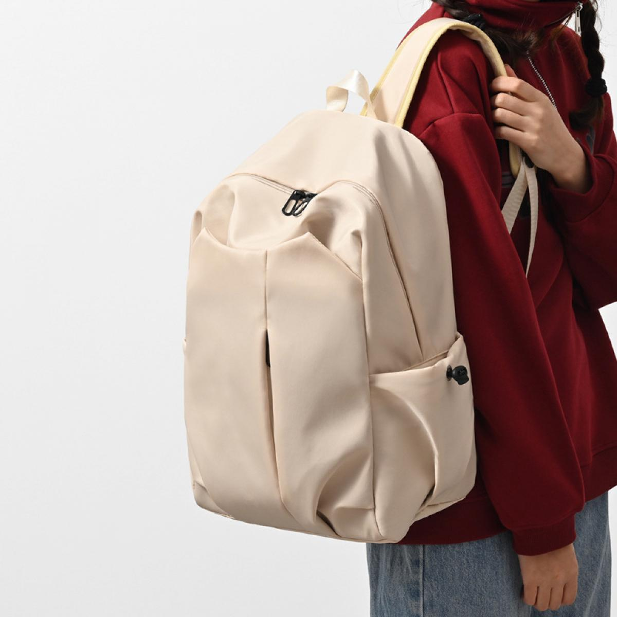 Eine Person mit einem beigefarbenen Rucksack und einem roten Kapuzenpullover. Der Rucksack hat schwarze Reißverschlüsse und Taschen. Die Person steht vor einem weißen Hintergrund.