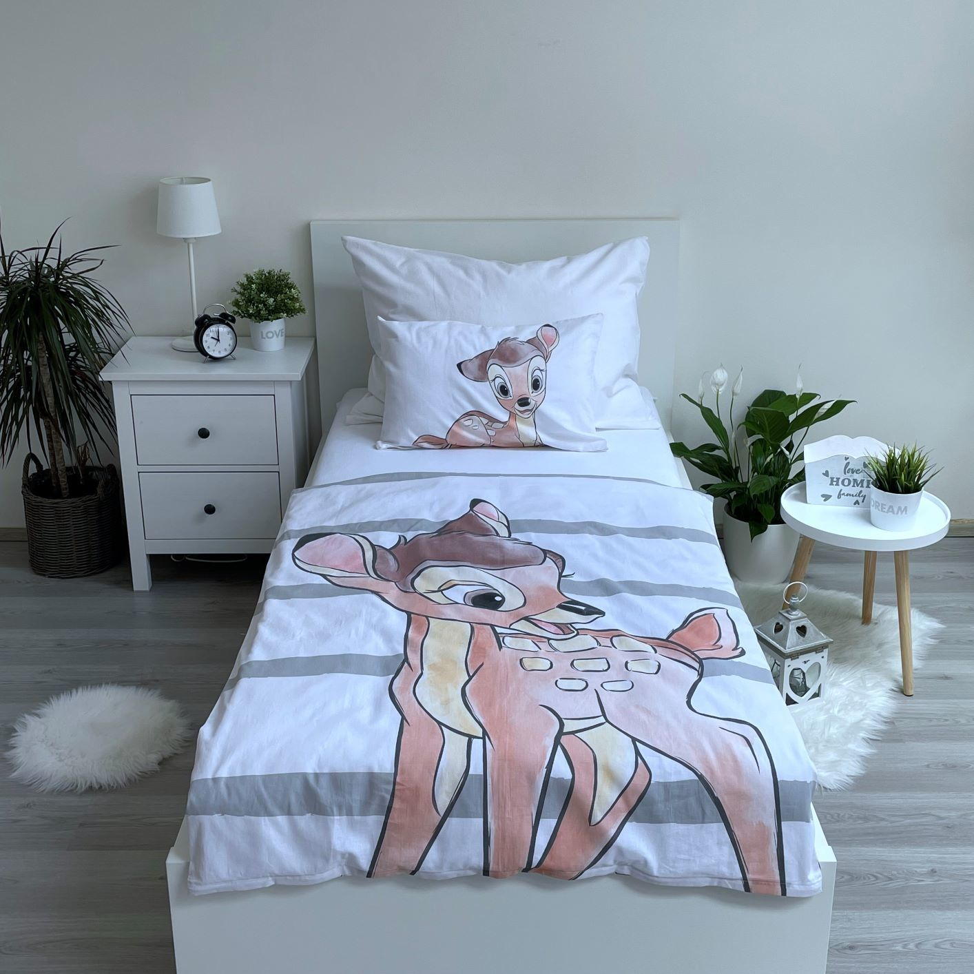 Ein Bett mit Bambi-Bettwäsche. Ein weißer Nachttisch hat eine Lampe und eine Pflanze. Ein kleiner Tisch hat eine Pflanze und ein Schild. Grau gestreifter Boden.