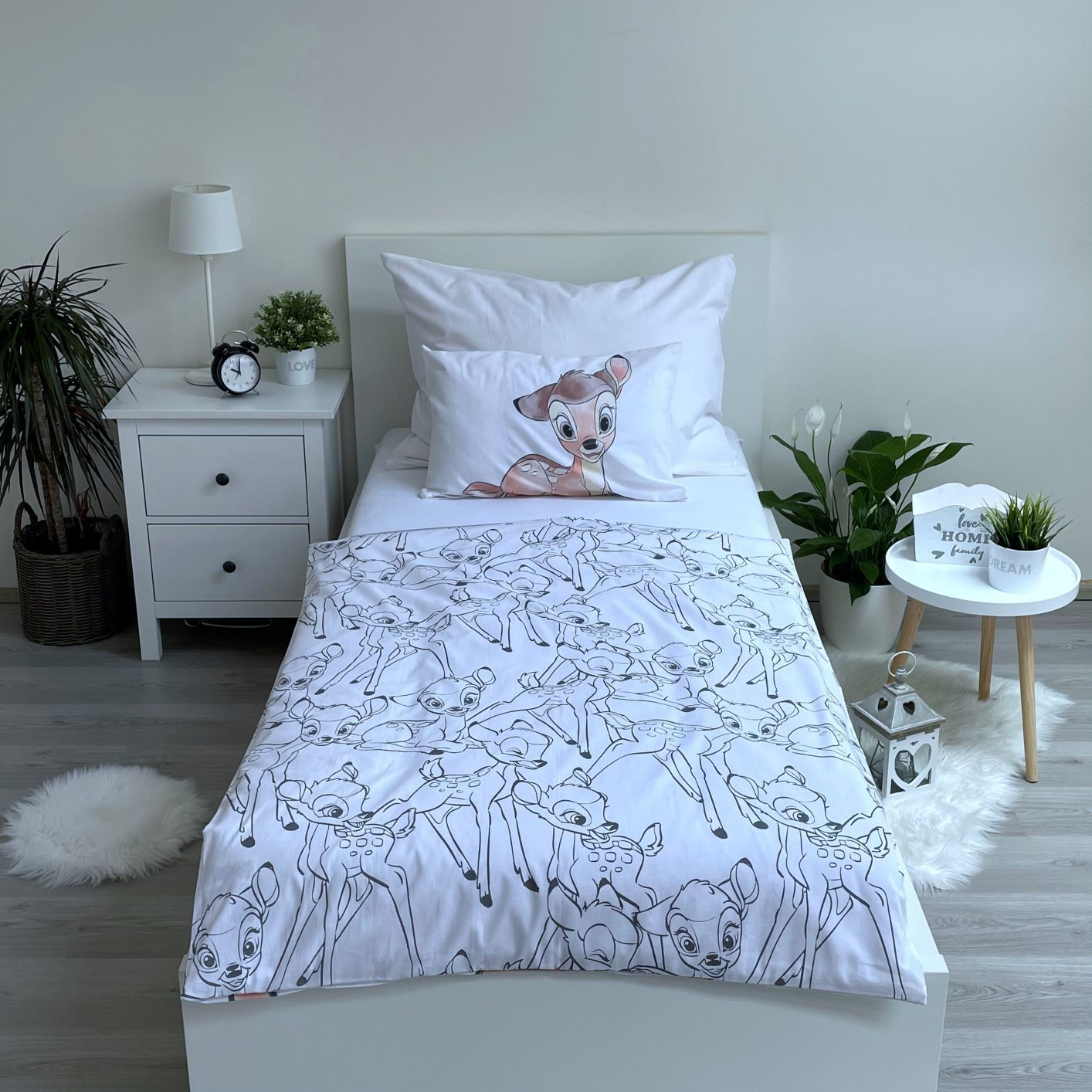 Ein Kinderbett mit Bettwäsche und Kissen im Bambi-Design. Ein Nachttisch mit Lampe und Pflanze. Eine weitere Pflanze und ein kleiner Tisch.