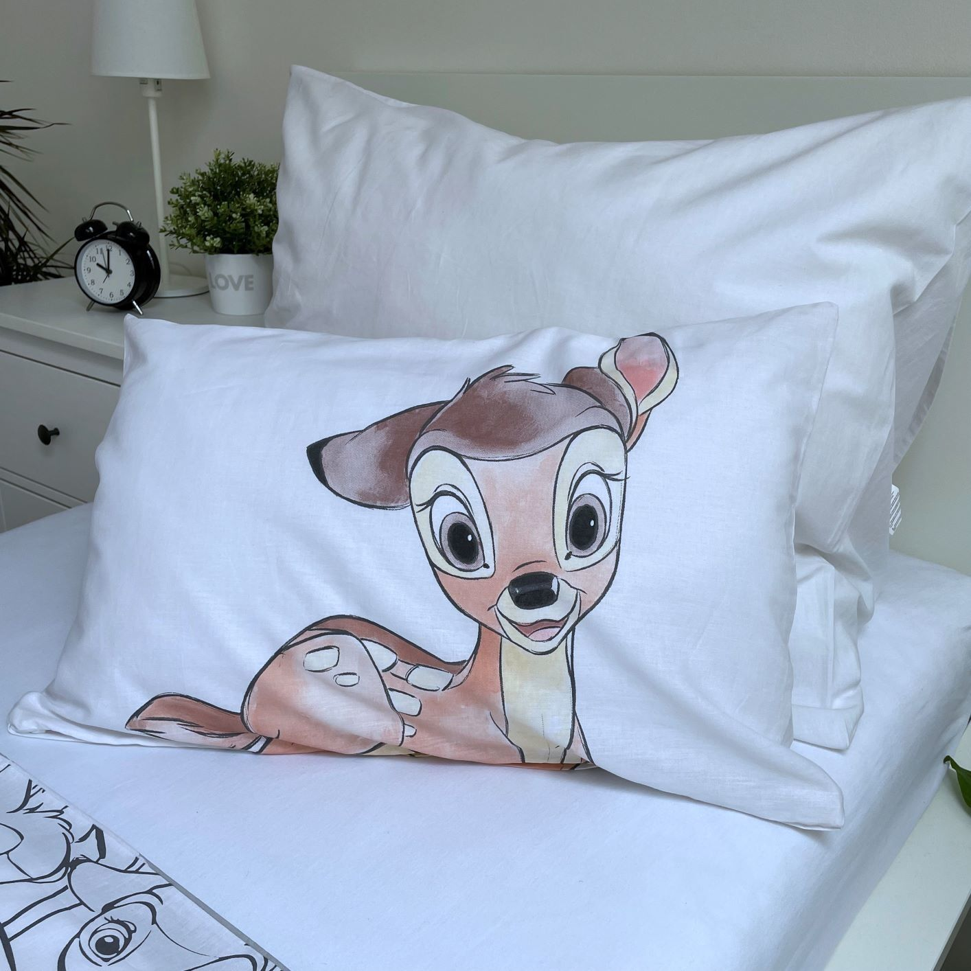 Ein weißes Kissen mit einer Bambi-Illustration liegt auf einem Bett mit weißen Laken. Dahinter steht ein Nachttisch mit Lampe und Wecker.