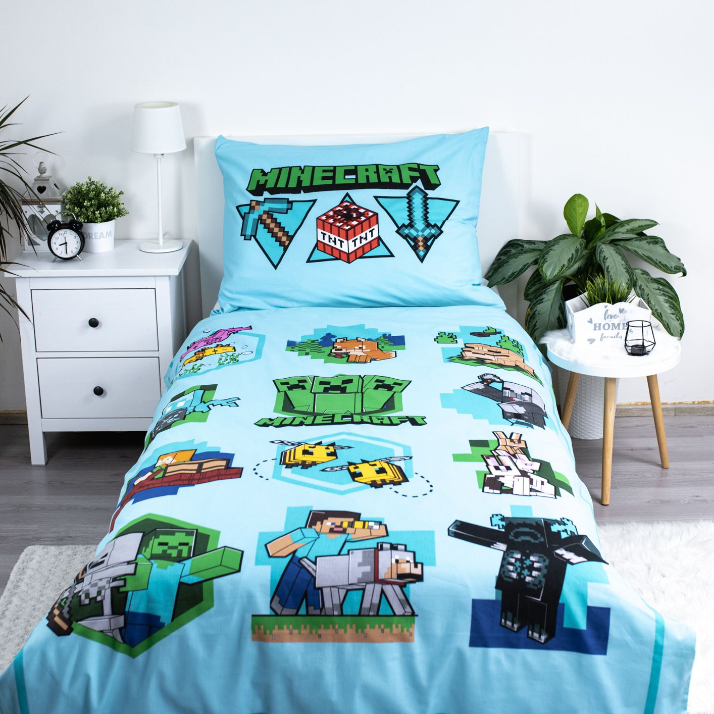 Ein Bett mit einem blauen Bettwäsche-Set im Minecraft-Design. Es zeigt verschiedene Spielcharaktere und Logos. Nachttisch und Pflanzen sind ebenfalls sichtbar.