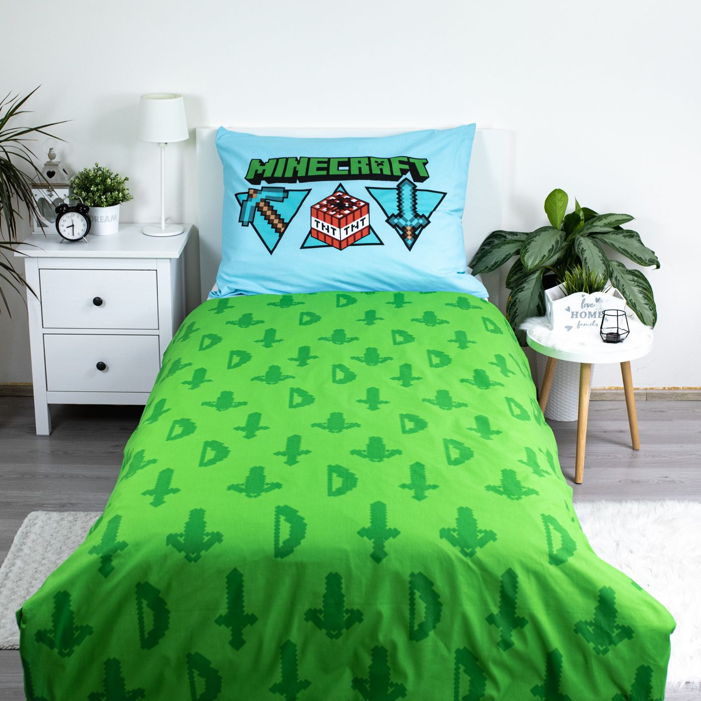 Ein grünes Minecraft-Bettwäsche-Set liegt auf einem Bett. Ein blaues Kissen mit dem Minecraft-Logo liegt darauf. Pflanzen und eine Lampe sind an den Seiten.