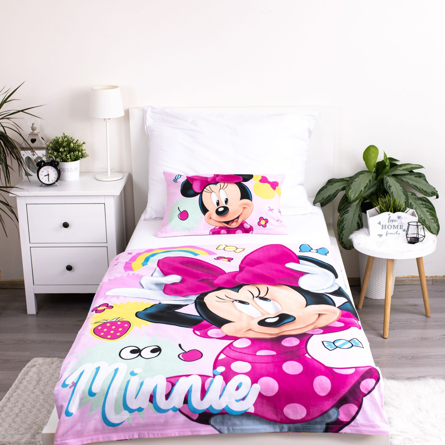 Bett mit Minnie-Maus-Bettwäsche. Weißer Nachttisch mit Lampe und Uhr. Pflanze rechts. Weiße Wand und Holzboden.