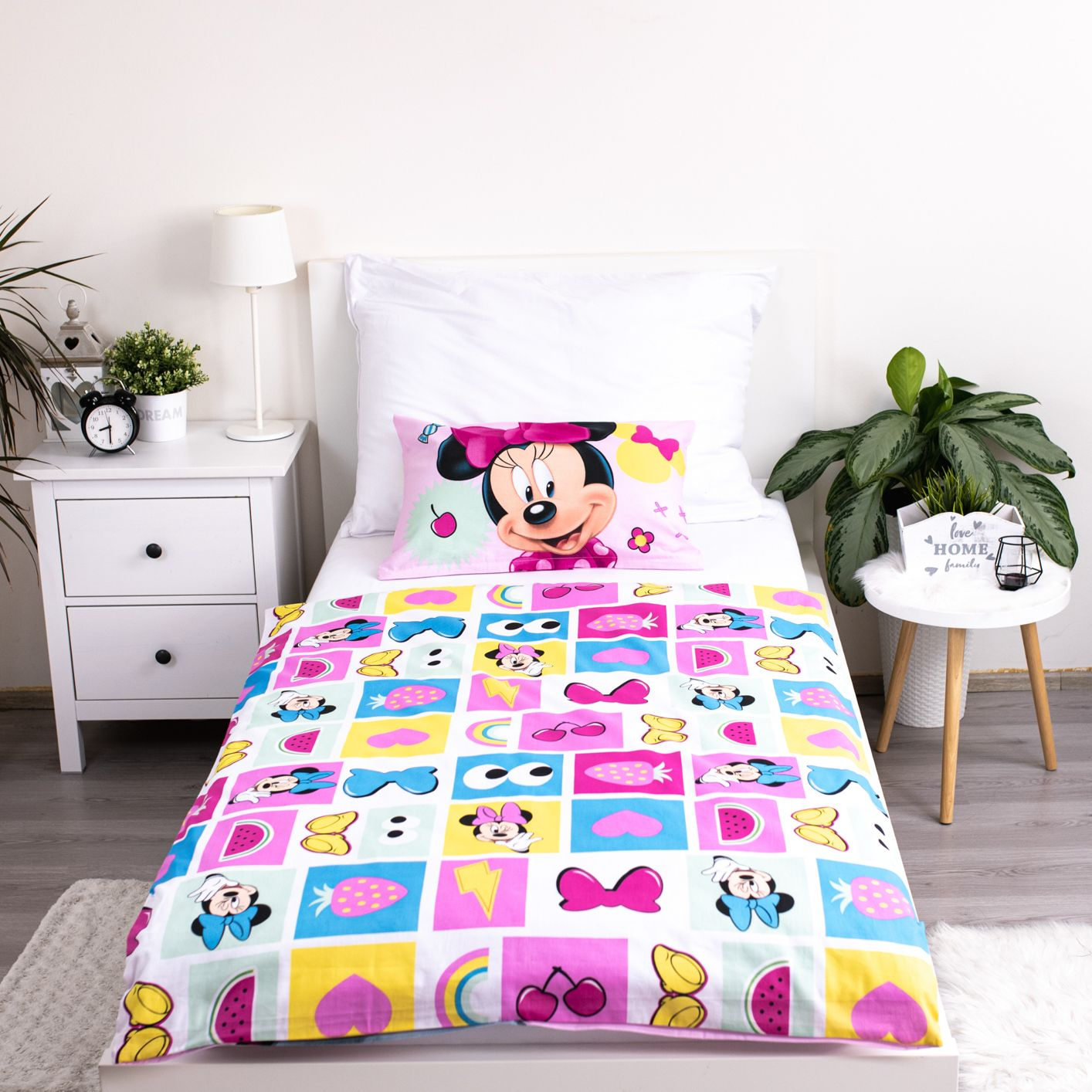 Ein Kinderbett mit Minnie Mouse-Bettwäsche. Ein weißer Nachttisch und ein kleiner Tisch mit Pflanzen. Auf dem Nachttisch steht eine weiße Lampe.