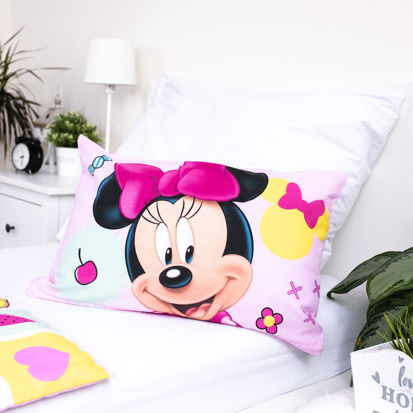 Ein pinkfarbenes Kissen mit Minnie Mouse auf einem weißen Bett. Eine Lampe, eine Uhr und Pflanzen sind im Hintergrund.