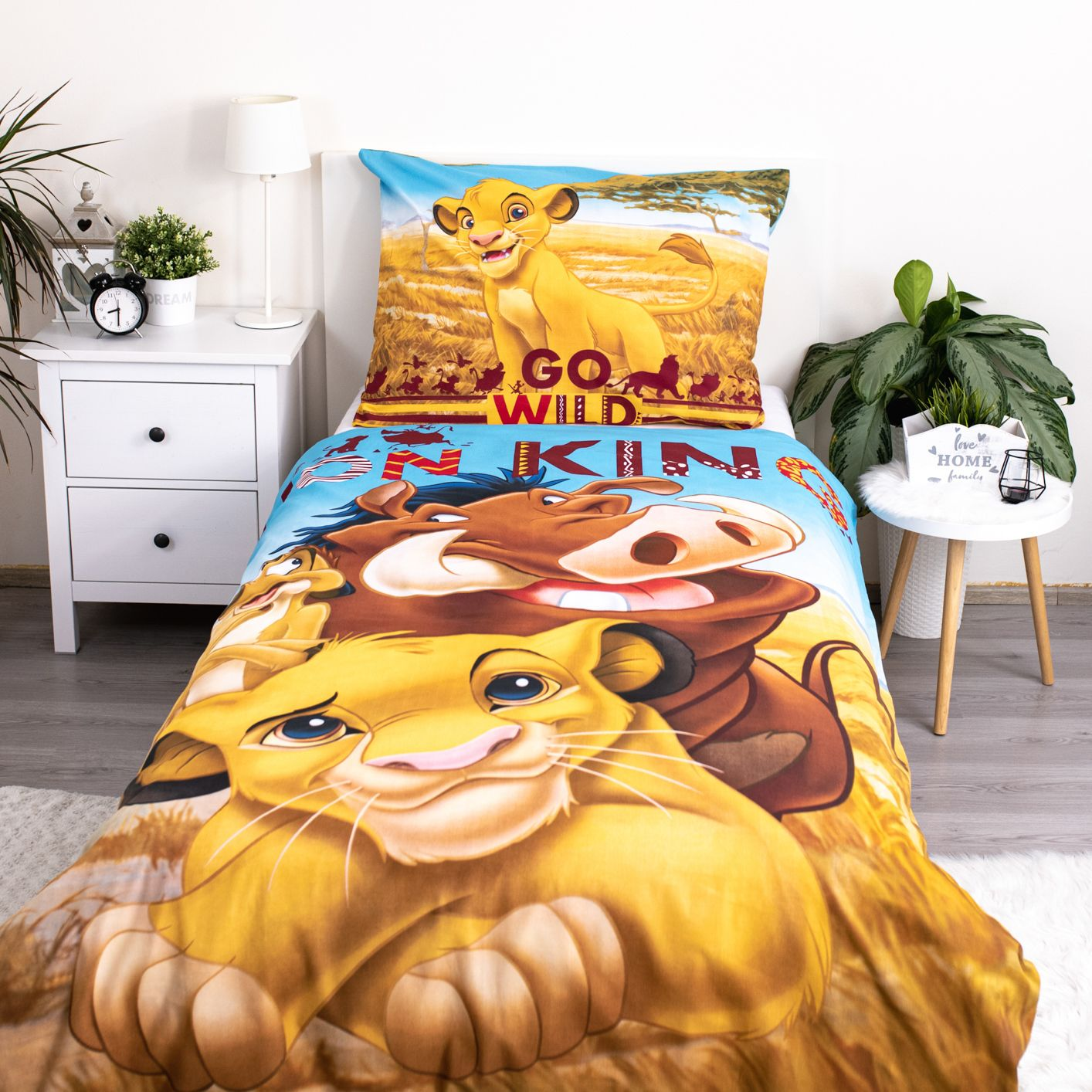 Lion King Bettwäsche auf einem weißen Bett. Simba, Pumbaa und Timon sind auf Bettdecke und Kissen abgebildet. Weißer Nachttisch und Pflanze.
