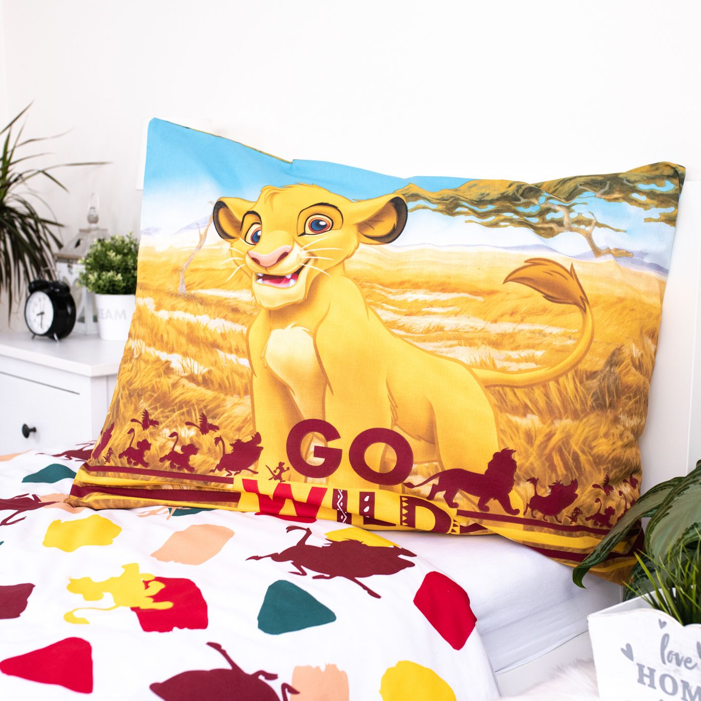 Ein Kissen mit Simba aus König der Löwen. Das Bett hat ein weißes Laken mit bunten Formen. Ein Nachttisch mit Pflanzen und einer Uhr ist sichtbar.