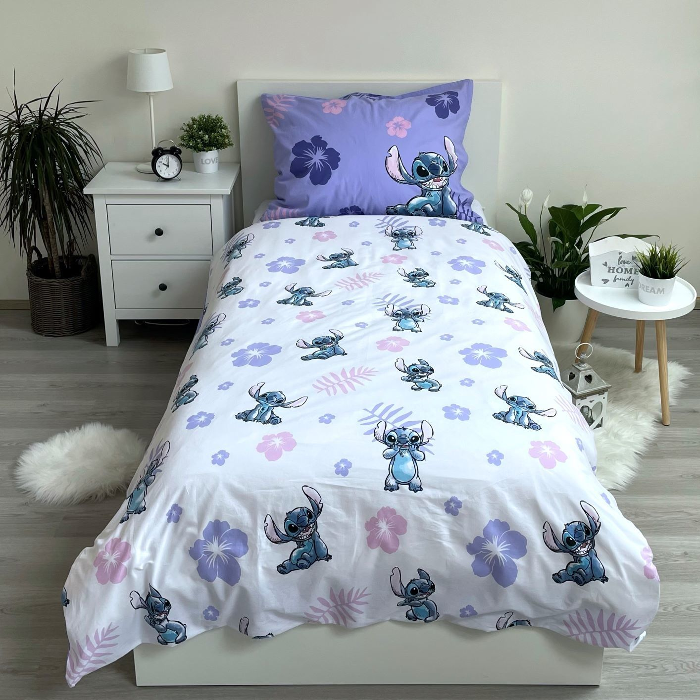 Ein Bett mit Stitch-Bettwäsche und Kissen. Neben dem Bett befindet sich ein Nachttisch mit Lampe, Uhr und Pflanzen.