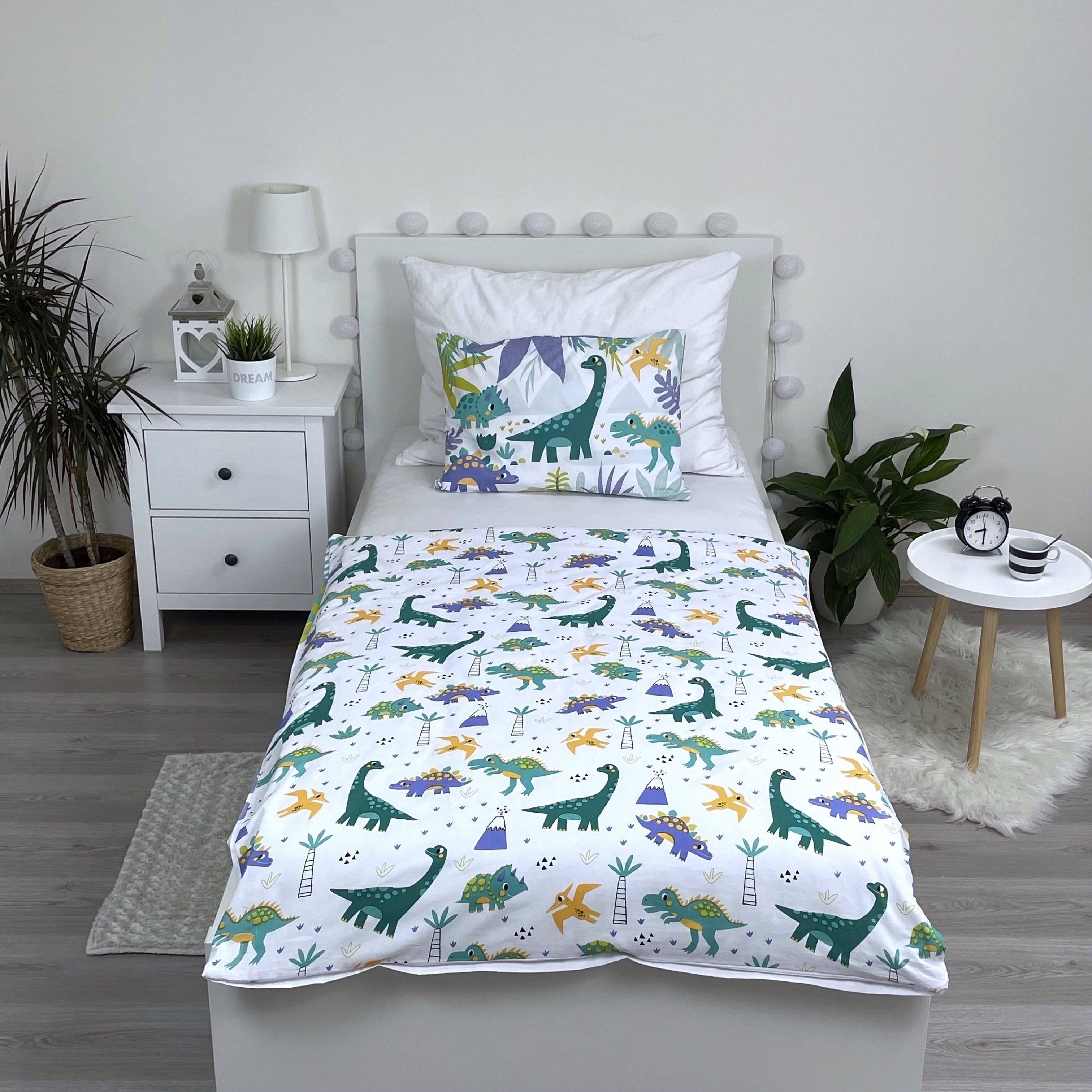 Ein Bett mit Dinosaurier-Bettwäsche, ein kleiner Tisch mit Uhr, ein weißer Nachttisch und eine Topfpflanze.