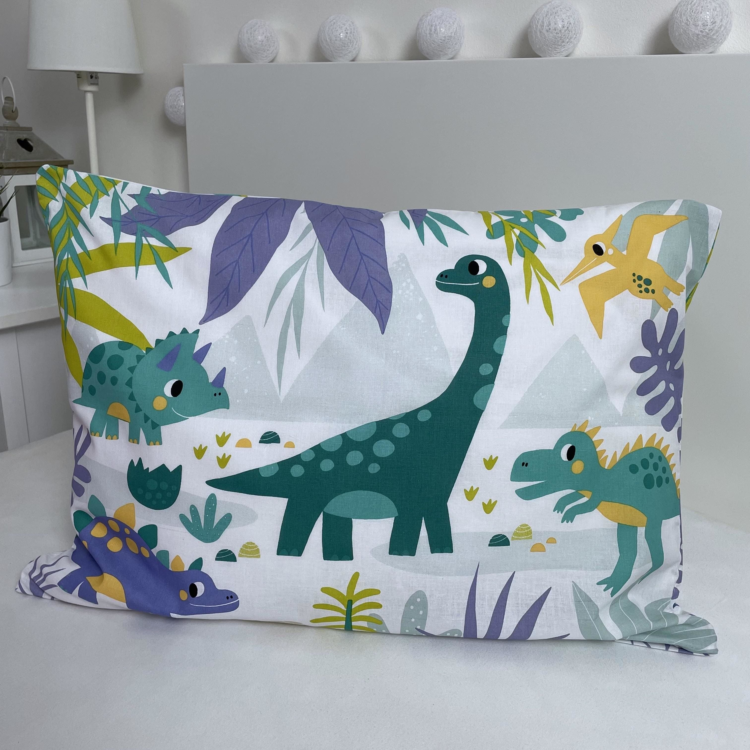 Kissen mit Dinosaurierdruck. Mit einem grünen Brachiosaurus, einem Triceratops, einem Pterodactylus und Blättern auf weißem Hintergrund.