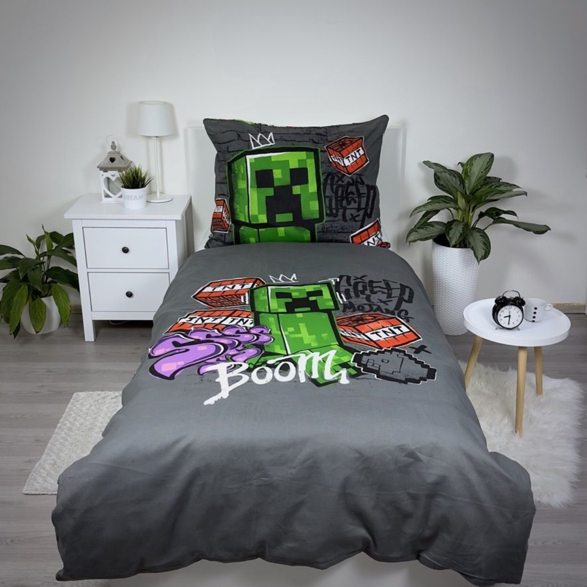 Ein Bett mit Minecraft-Bettwäsche. Ein grüner Creeper befindet sich auf der Bettdecke. Es gibt einen weißen Nachttisch, eine Pflanze und einen kleinen Tisch.